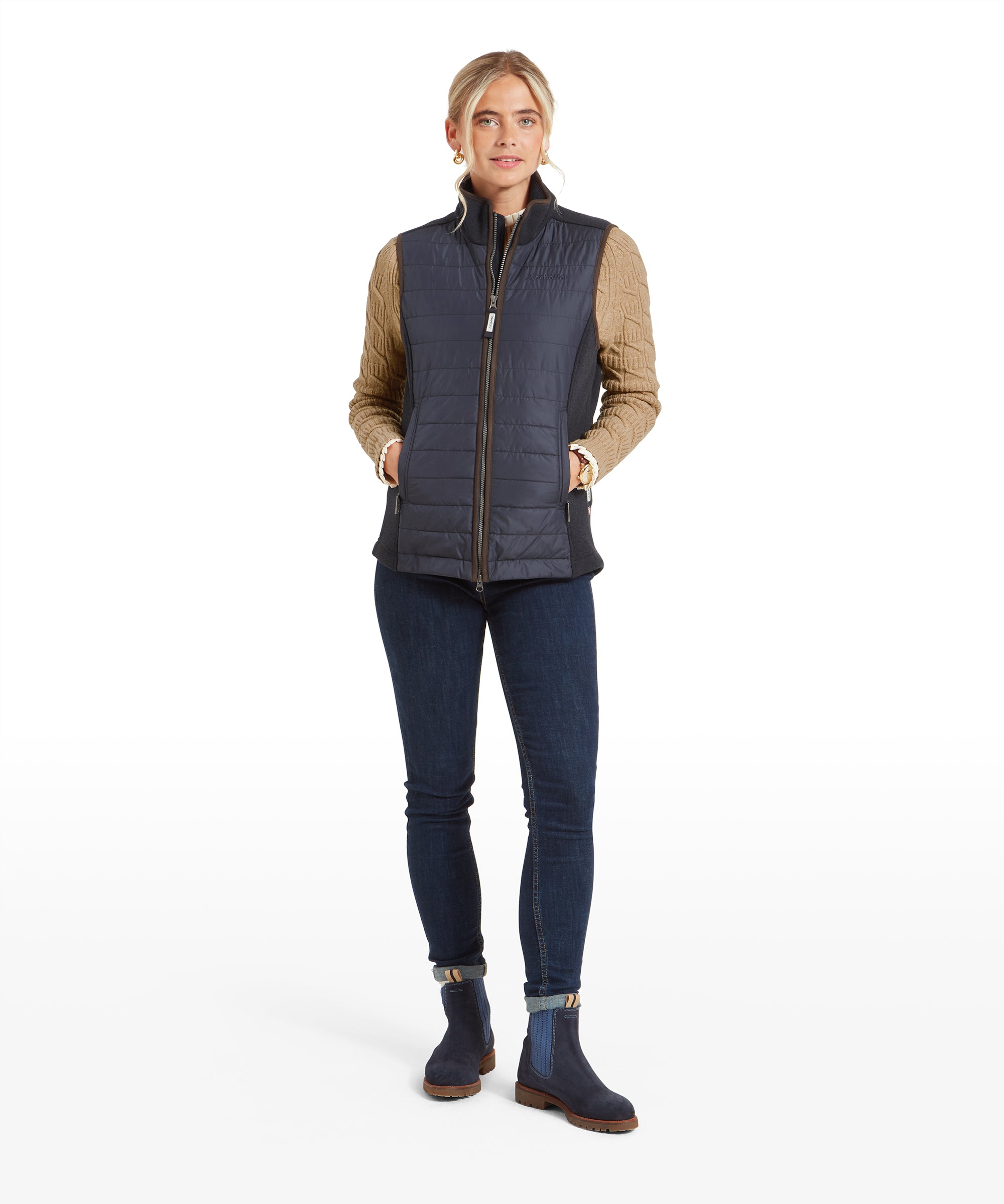 The same woman is standing in a full-body view, with her hands in the pockets of the Schöffel Lyndon Hybrid Gilet for Women in Blue. She is wearing skinny jeans and navy boots with light blue elastic sides.