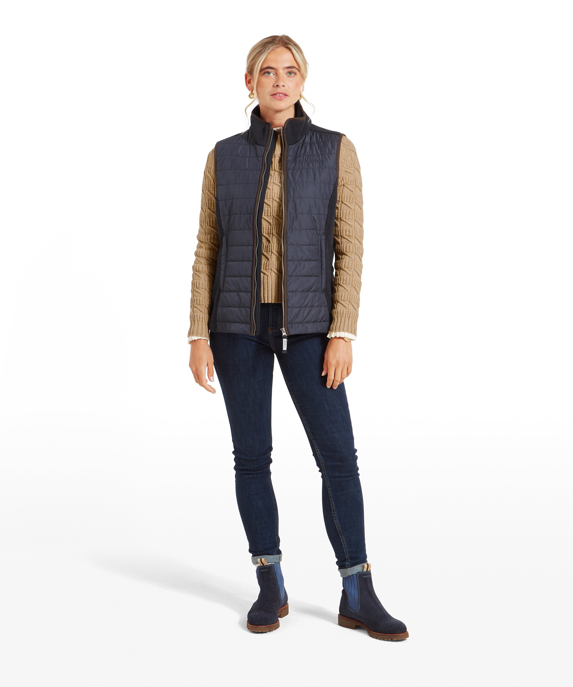 The woman standing in a full-body view, hands in her pockets, wearing the Schöffel Lyndon Hybrid Gilet for Women in Blue, skinny jeans, and navy boots.