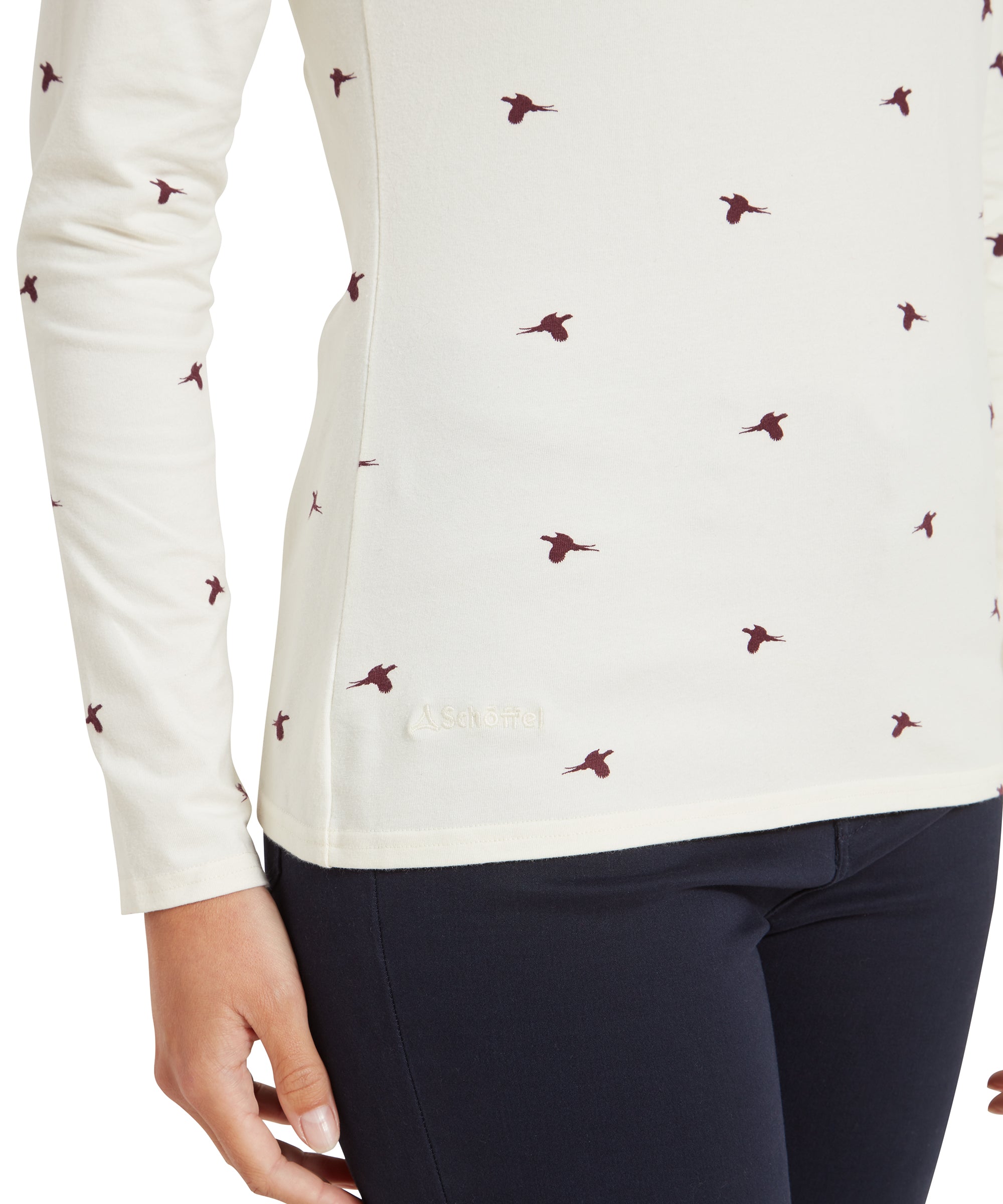 A close-up of the side of a Schöffel Malham Top for Women in Red featuring small maroon bird prints and the embroidered Schöffel logo near the hem. The image also shows part of the dark navy pants the woman is wearing.