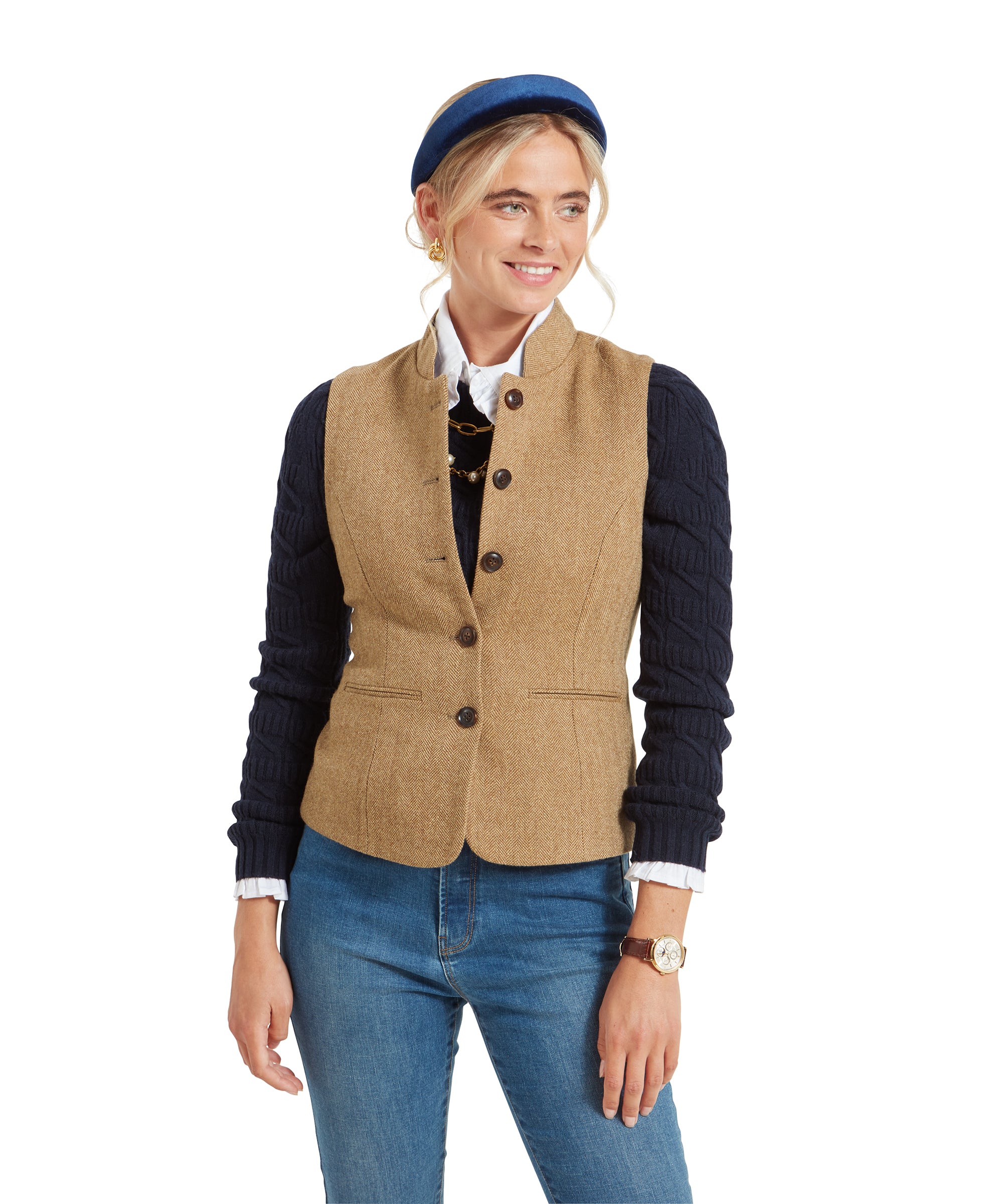 A woman wearing a Schöffel Millport Tweed Nehru Waistcoat for Womens in Light Brown over a navy sweater and white shirt. She is also wearing blue jeans and a blue headband, smiling slightly.