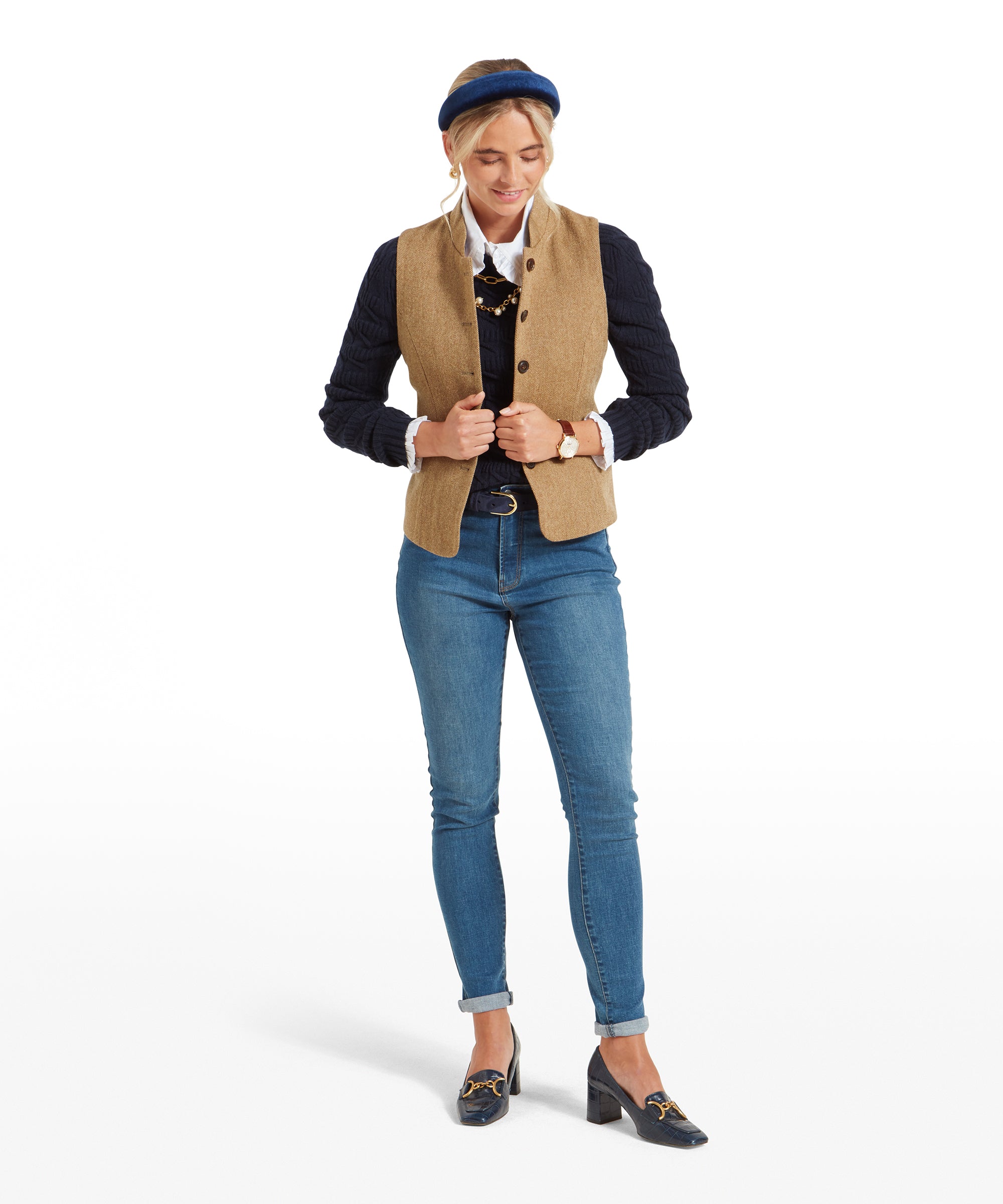 The same woman standing and adjusting her Schöffel Millport Tweed Nehru Waistcoat for Womens in Light Brown while looking down. She is dressed in a navy sweater, white shirt, and blue jeans.