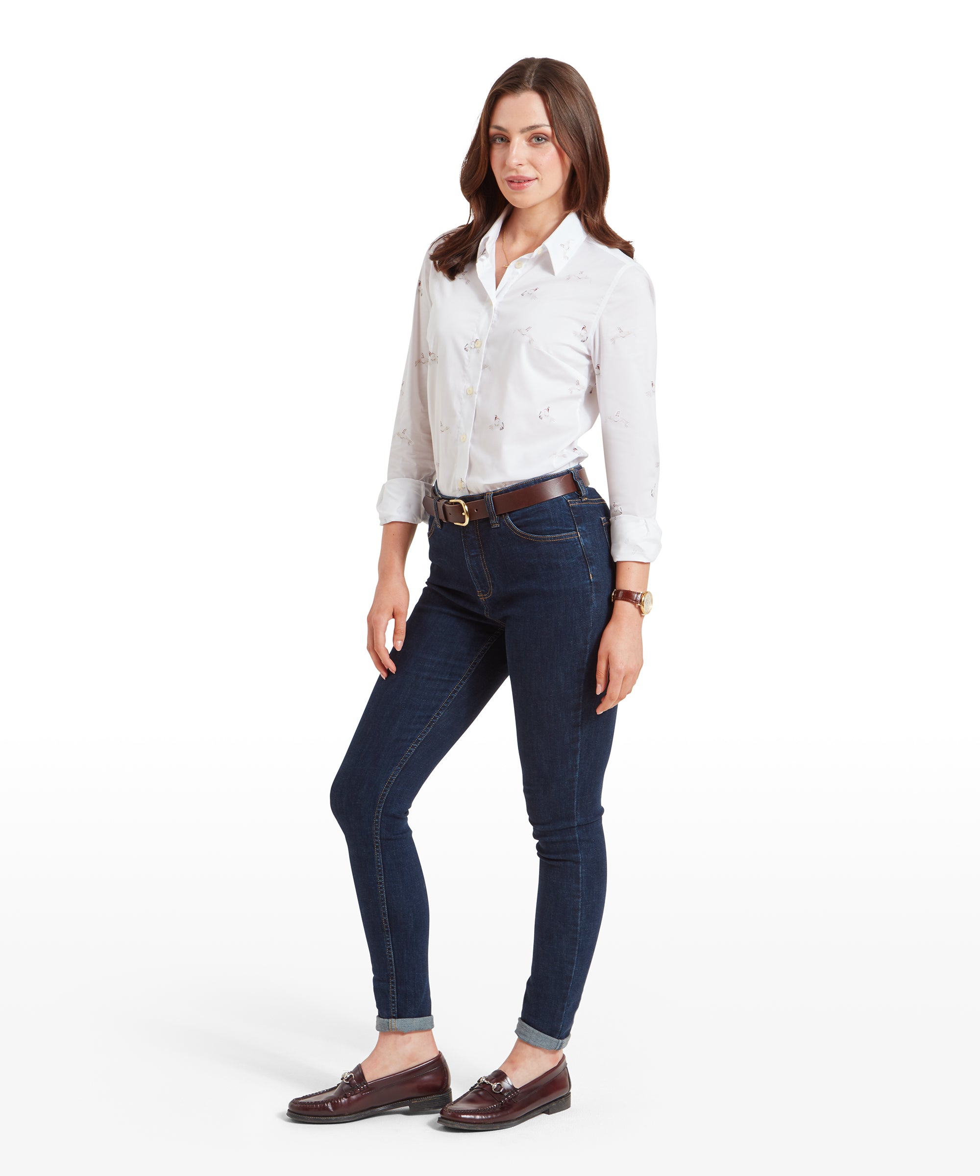 A full-body view of the woman wearing the Schöffel Norfolk Shirt for Womens in White Grey with a pheasant print, tucked into dark blue jeans. She is also wearing brown leather loafers and has her hands relaxed by her sides. Her hair is styled in soft waves, and she is smiling confidently.