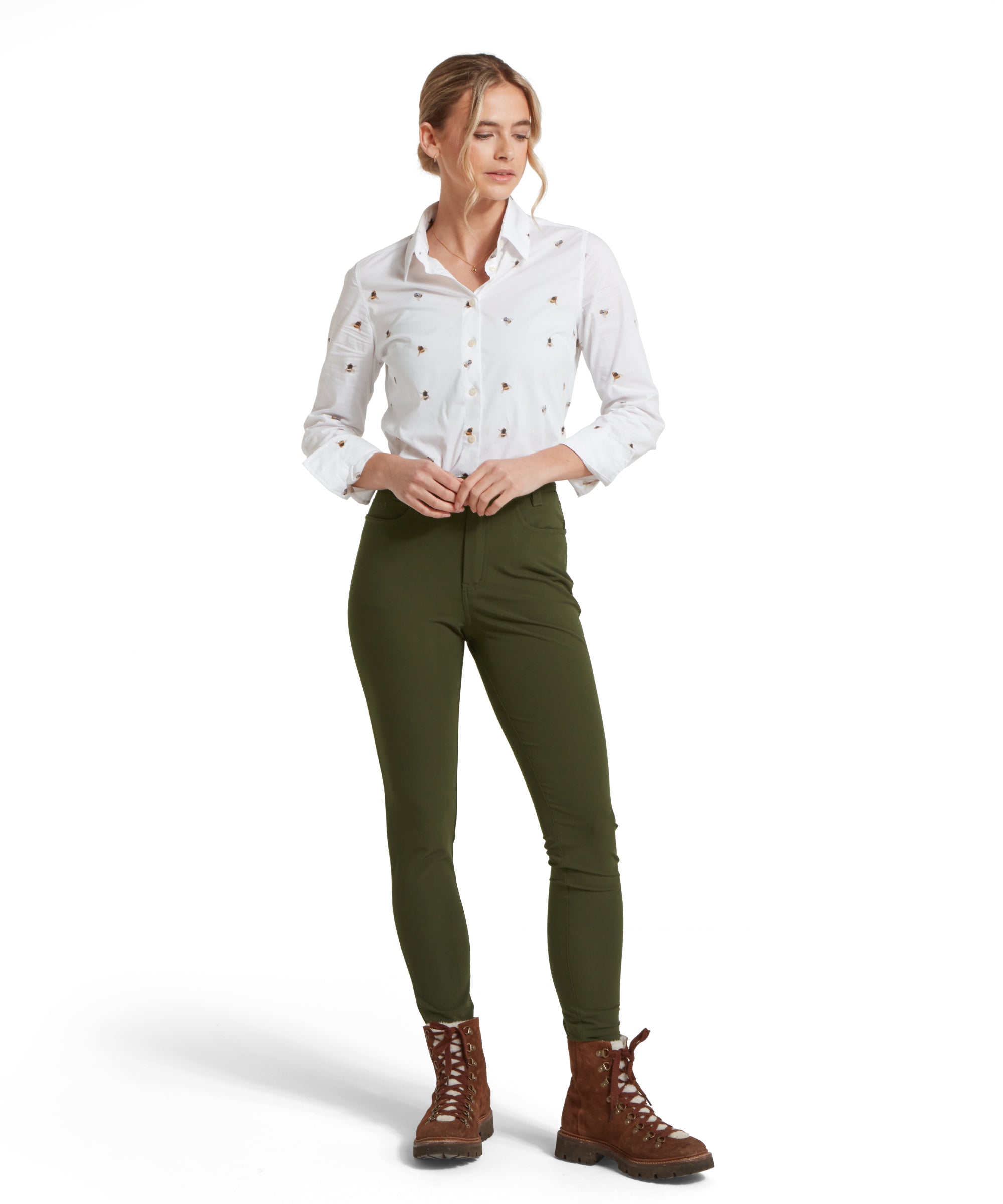 Full height image of a female model wearing a white Schöffel shirt with a bee pattern, paired with mid green trousers and brown boots.