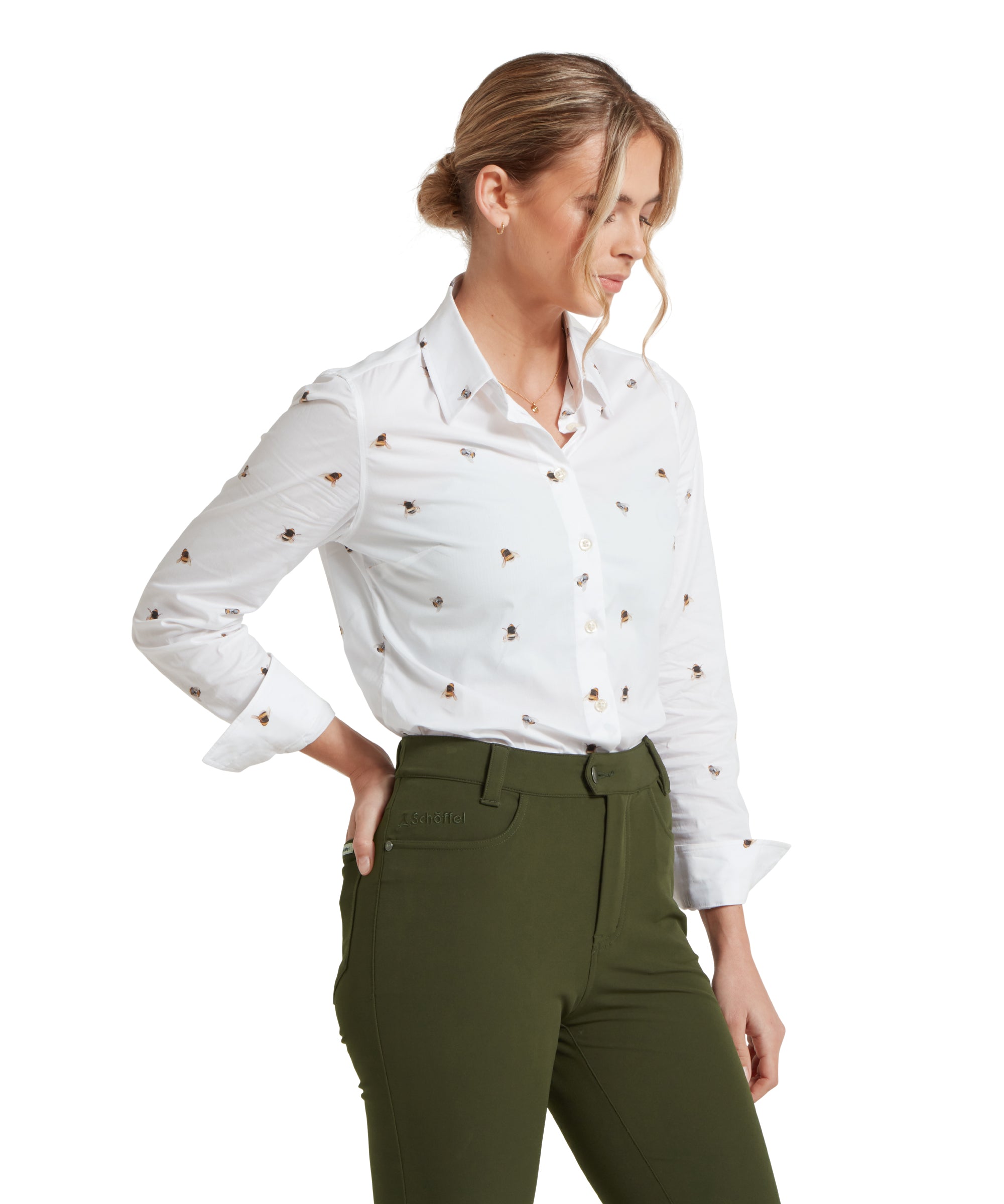 Side profile image of a female model wearing a white Schöffel shirt with a bee pattern, paired with mid green trousers. Model is standing in a relaxed pose with one hand on her hip and she is gazing down.