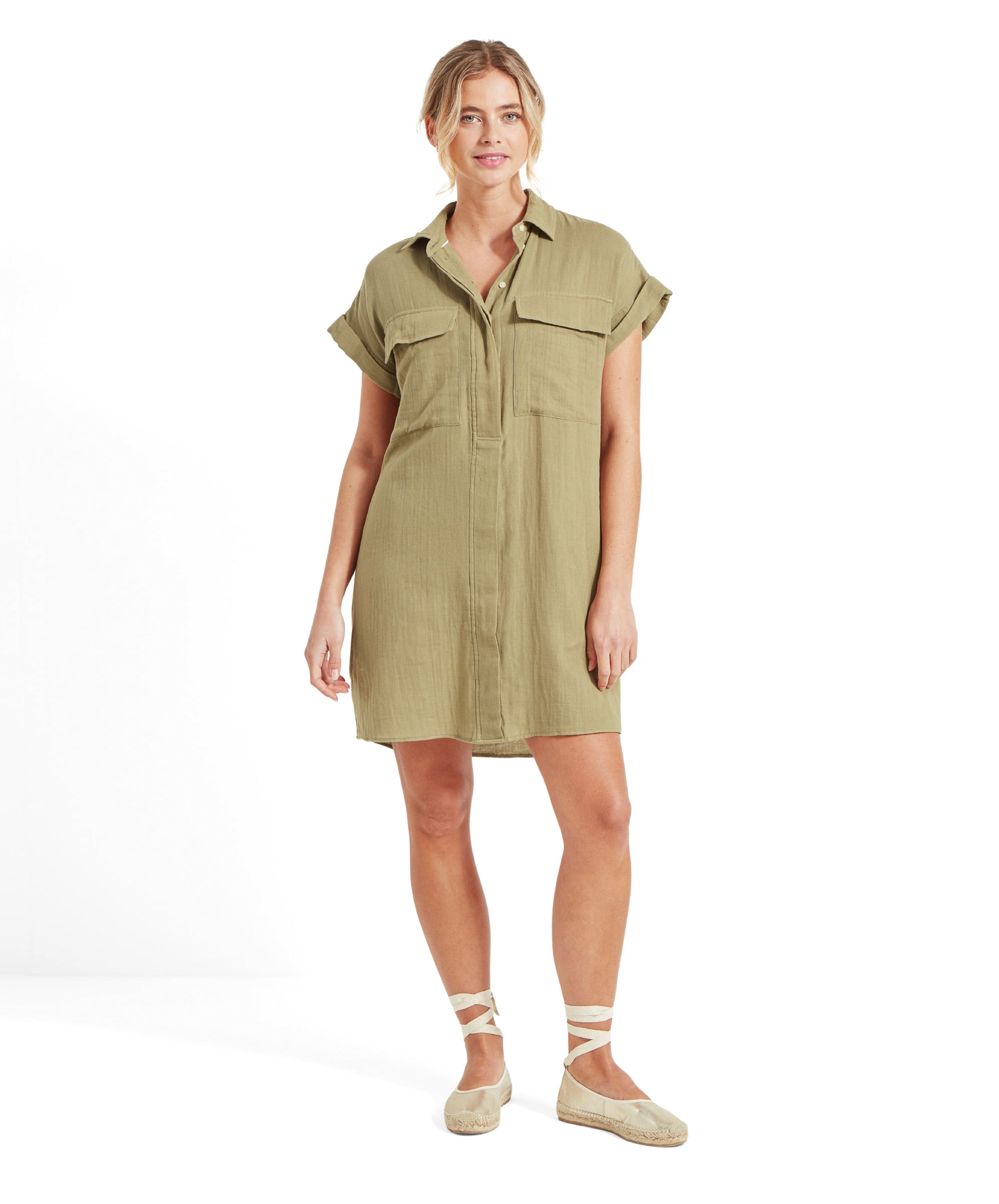 The same woman is standing and looking at the camera. She is wearing the same Schöffel Olive Dress for Women in Green with front pockets and a button-up design, along with beige espadrilles with ankle straps.