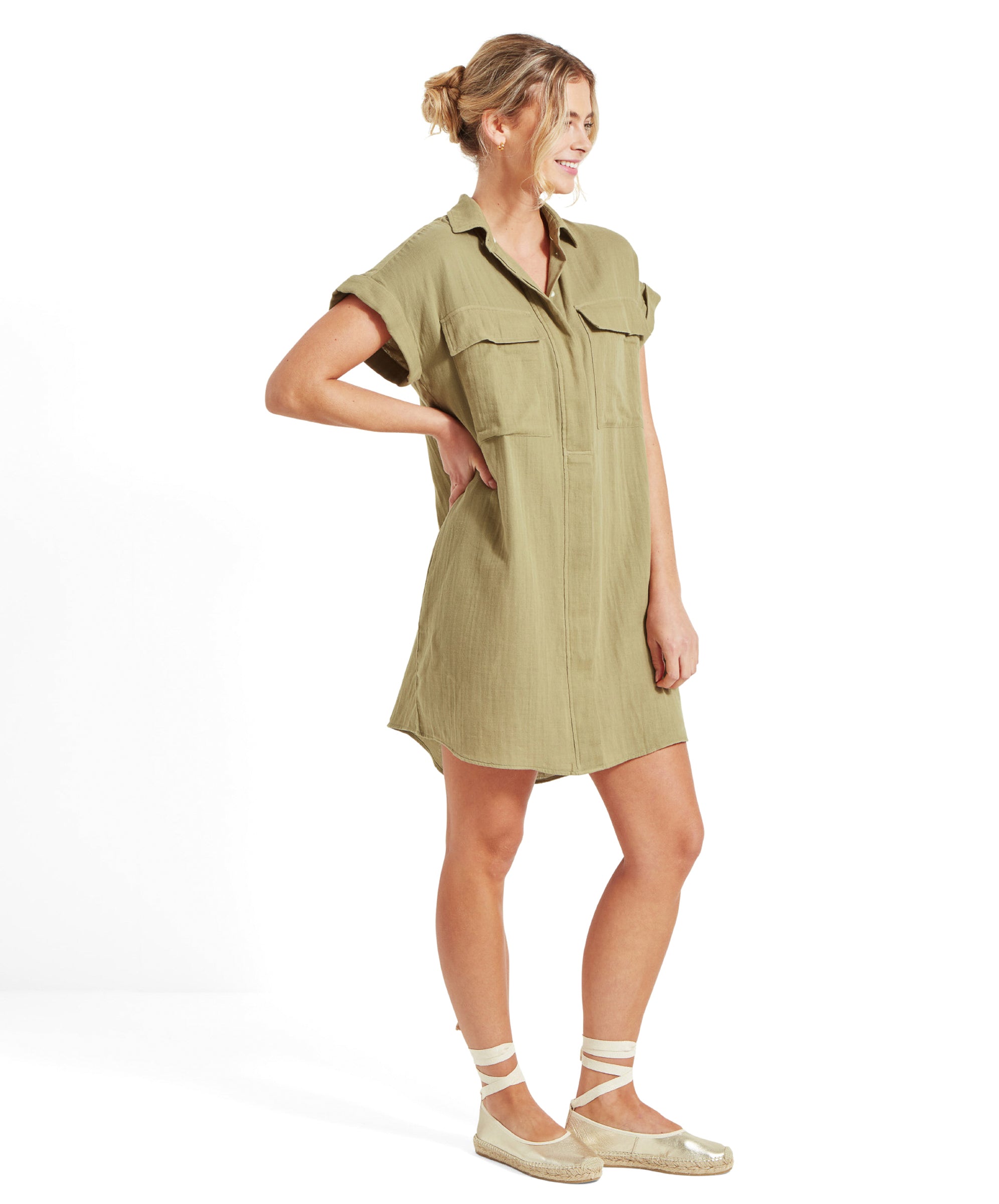 The woman is standing with one hand on her hip and smiling, wearing the Schöffel Olive Dress for Women in Green with front pockets and a button-up design. She is also wearing beige espadrilles with ankle straps.