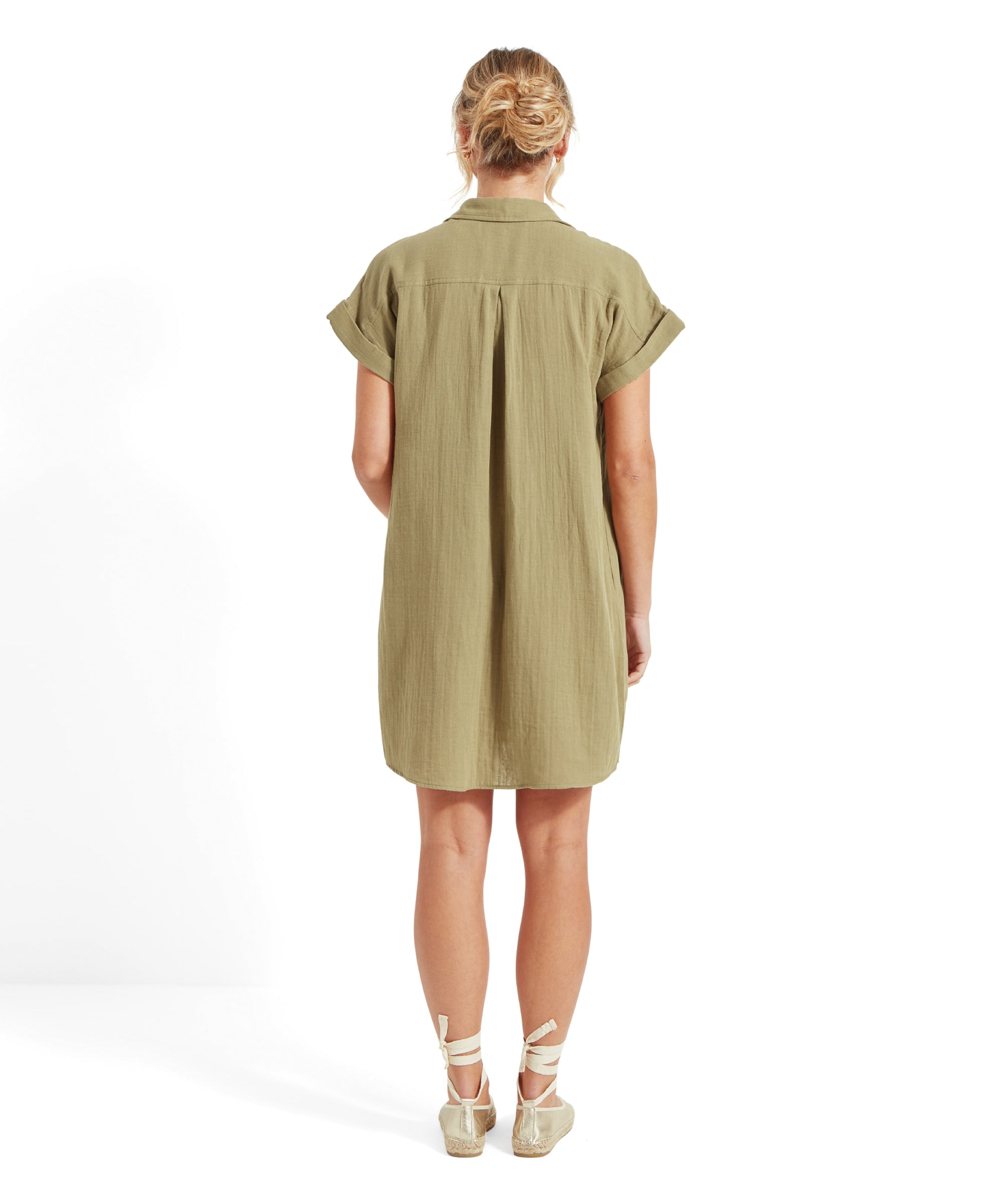 The woman is turned around, displaying the back of the Schöffel Olive Dress for Women in Green with a pleat detail. She is wearing beige espadrilles with ankle straps and has her hair tied back in a bun.