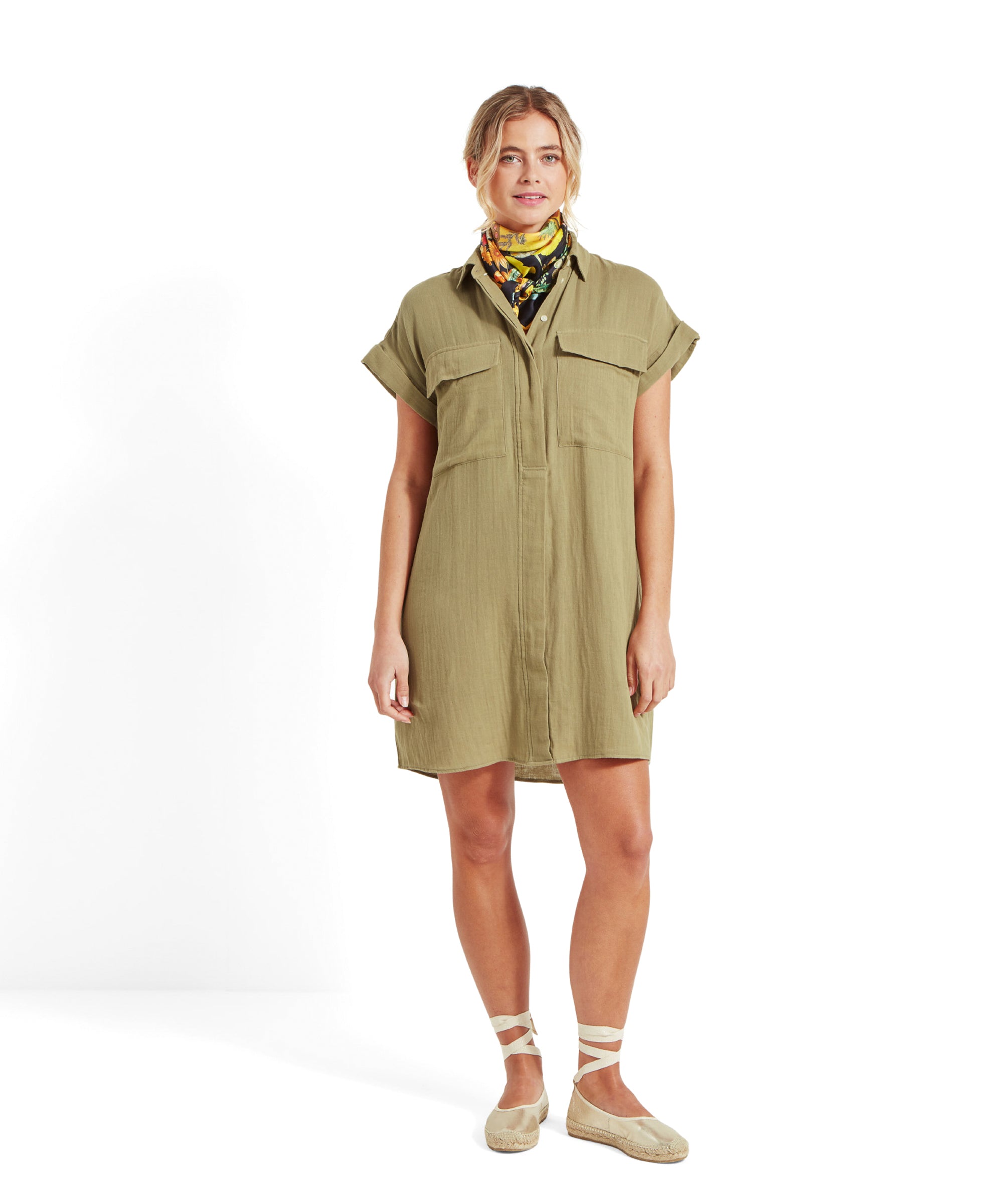 The woman is standing and smiling, wearing the Schöffel Olive Dress for Women in Green with front pockets and a button-up design. She is also wearing a colorful floral scarf around her neck and beige espadrilles with ankle straps.