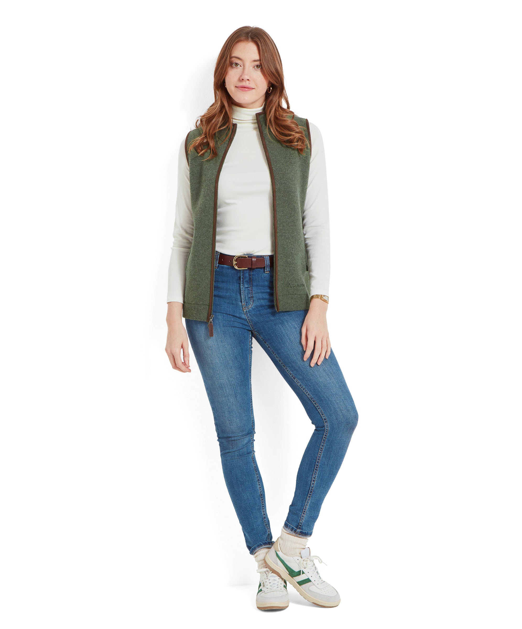 Full-body view of a woman wearing a Schöffel Women's Orkney Merino Gilet for Women in Green, white turtleneck, blue jeans, and white trainers, standing and looking at the camera.