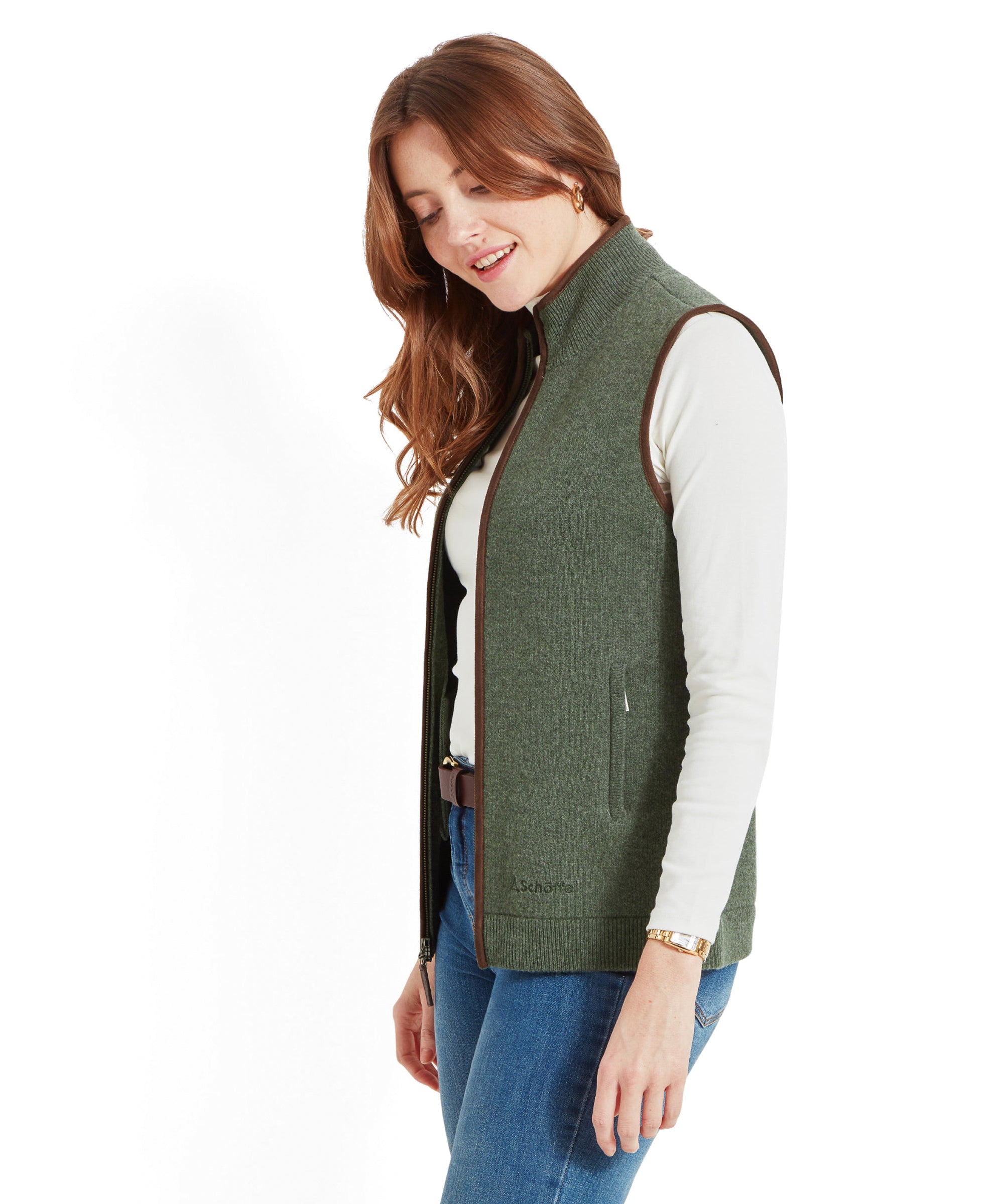 Side view of a woman wearing a Schöffel Women's Orkney Merino Gilet for Women in Green, white turtleneck, and blue jeans, looking down and smiling.