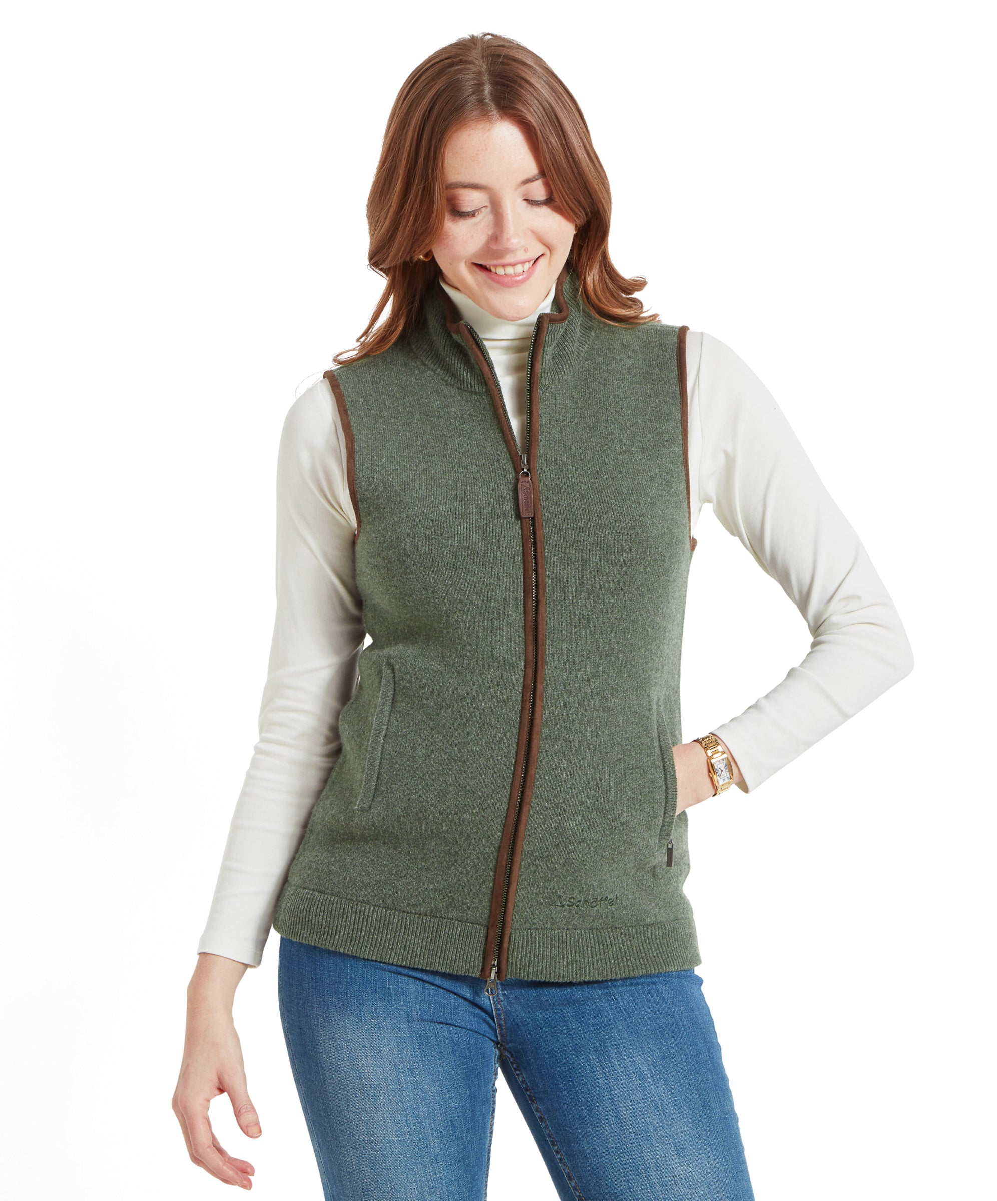 A woman wearing a Schöffel Women's Orkney Merino Gilet for Women in Green, white turtleneck, and blue jeans, smiling and looking down.