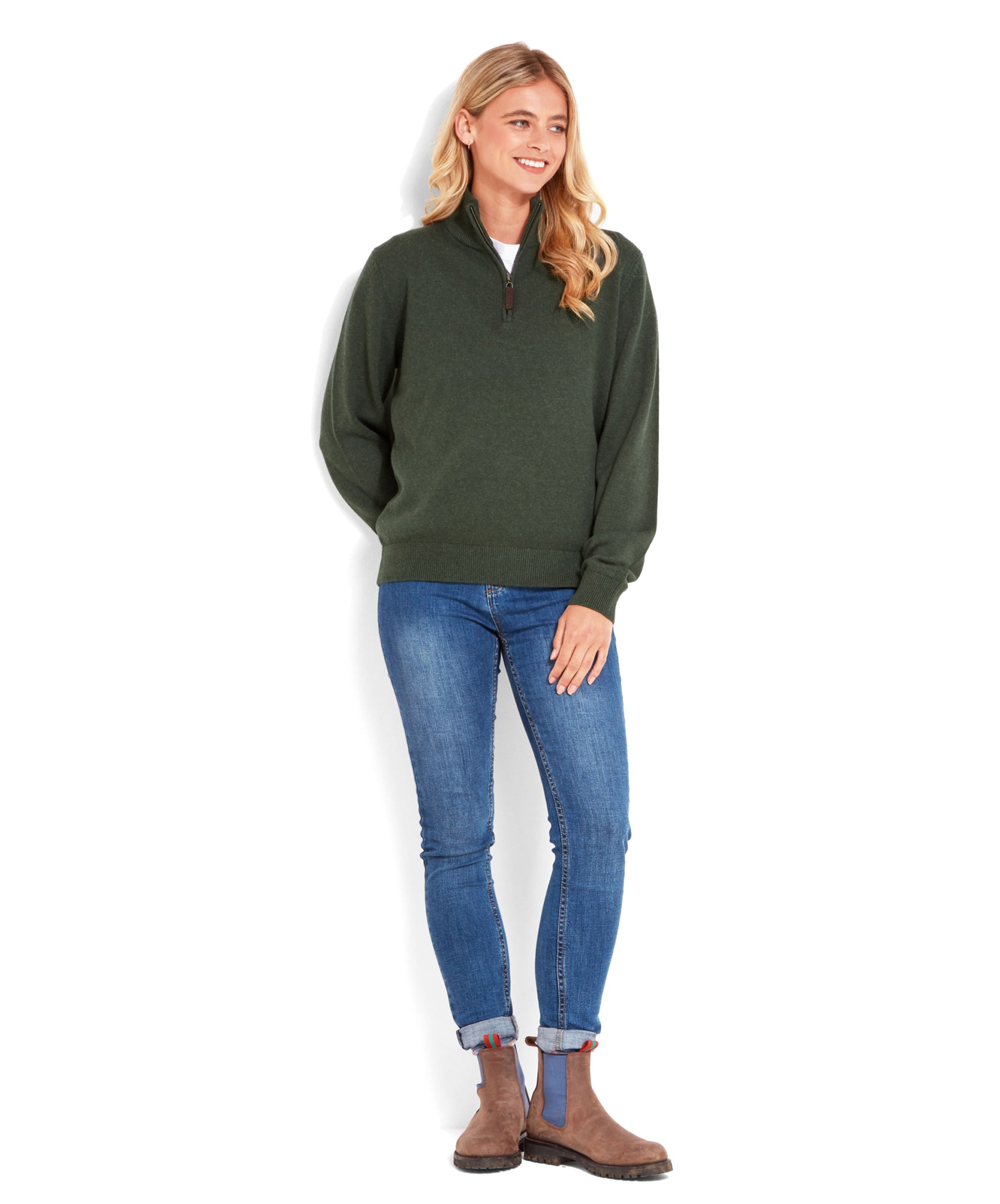 The same woman is standing and looking to the side, wearing the Schöffel Pateley Jumper for Women in Green and blue jeans. She has her hands in her pockets and is wearing brown boots.