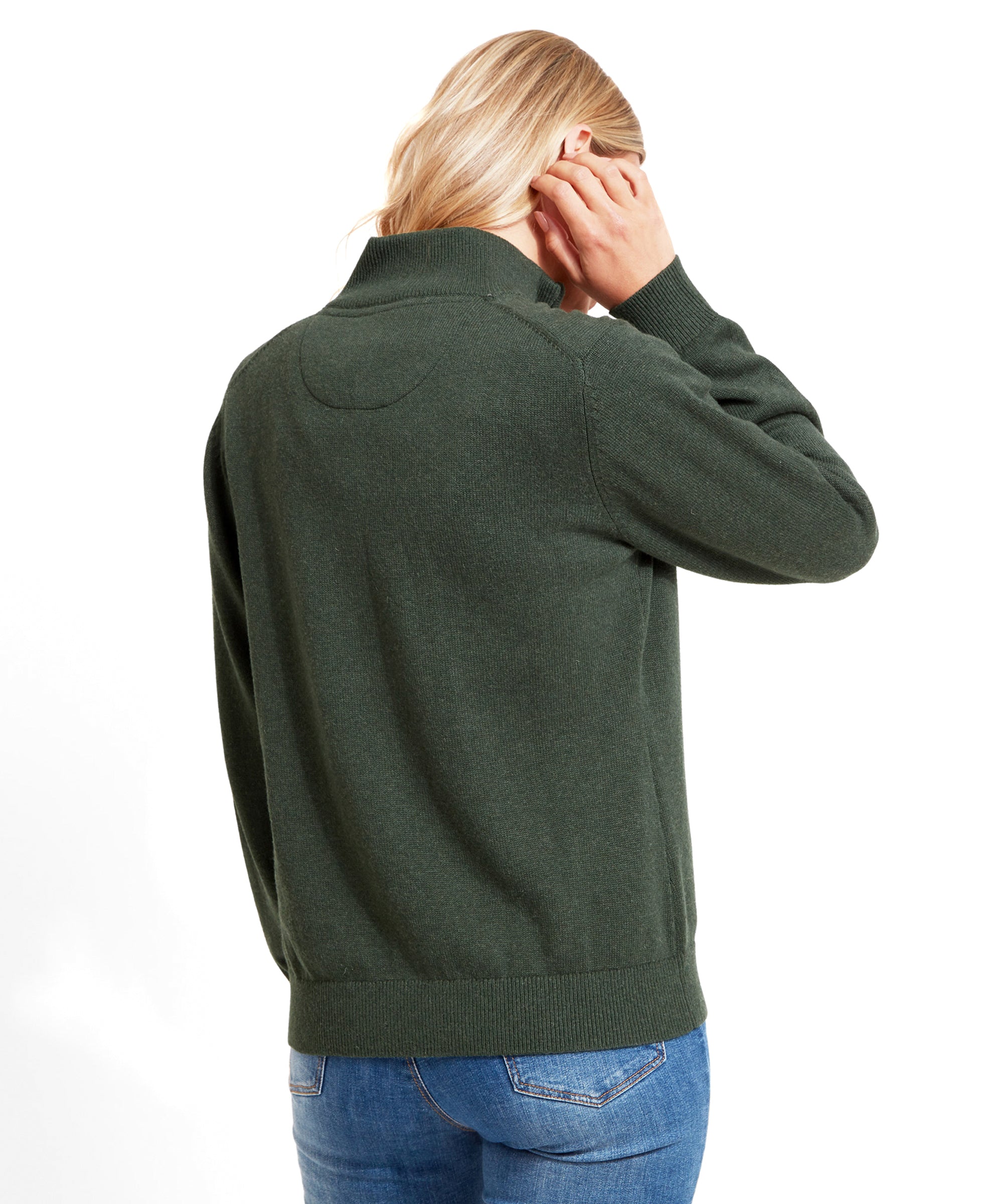 The woman is turned around, displaying the back of the Schöffel Pateley Jumper for Women in Green. She is adjusting her hair with one hand and wearing blue jeans.