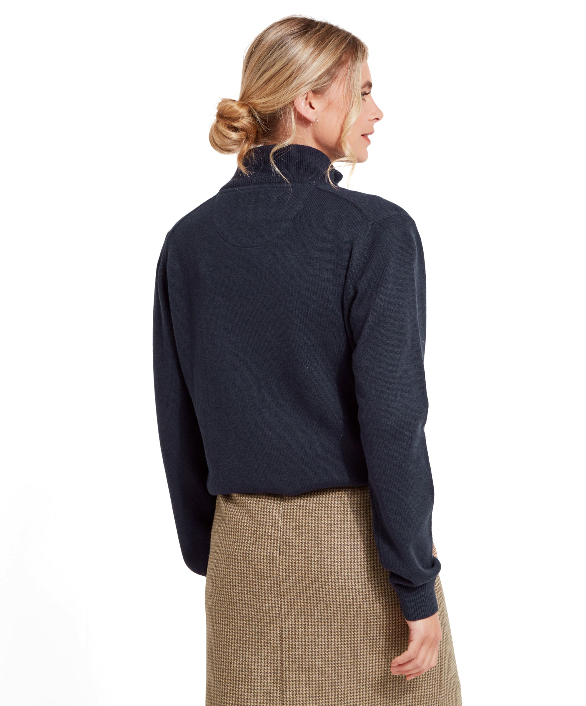 The woman is turned around, displaying the back of the Schöffel Pateley Jumper for Women in Navy. She is wearing a brown checkered skirt and has her hair tied back in a bun.