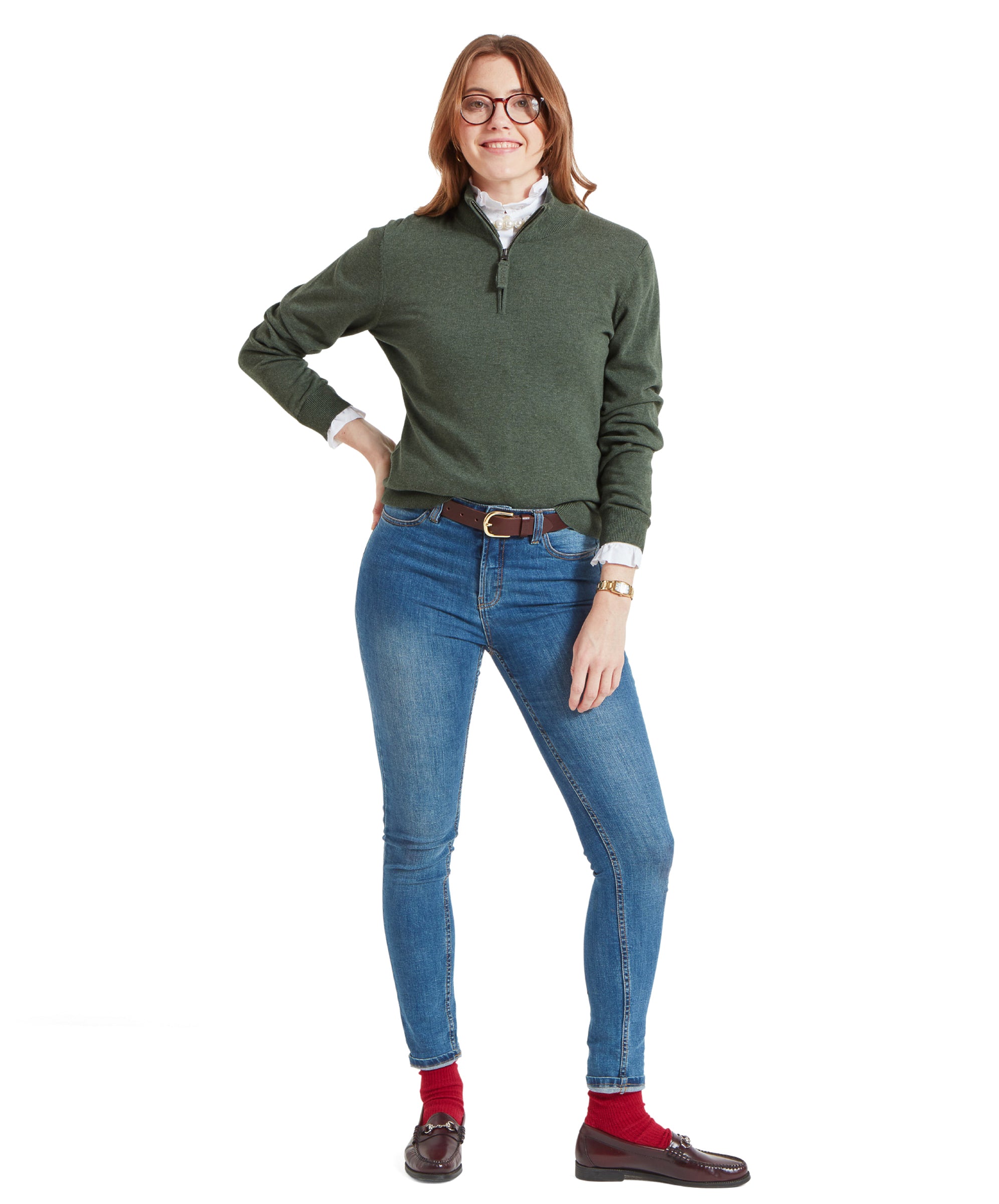 Full-body shot of the woman in the Schöffel Polperro Pima Cotton 1/4 Zip for Women in Green, blue jeans, red socks, and brown loafers, standing with one hand on her hip.