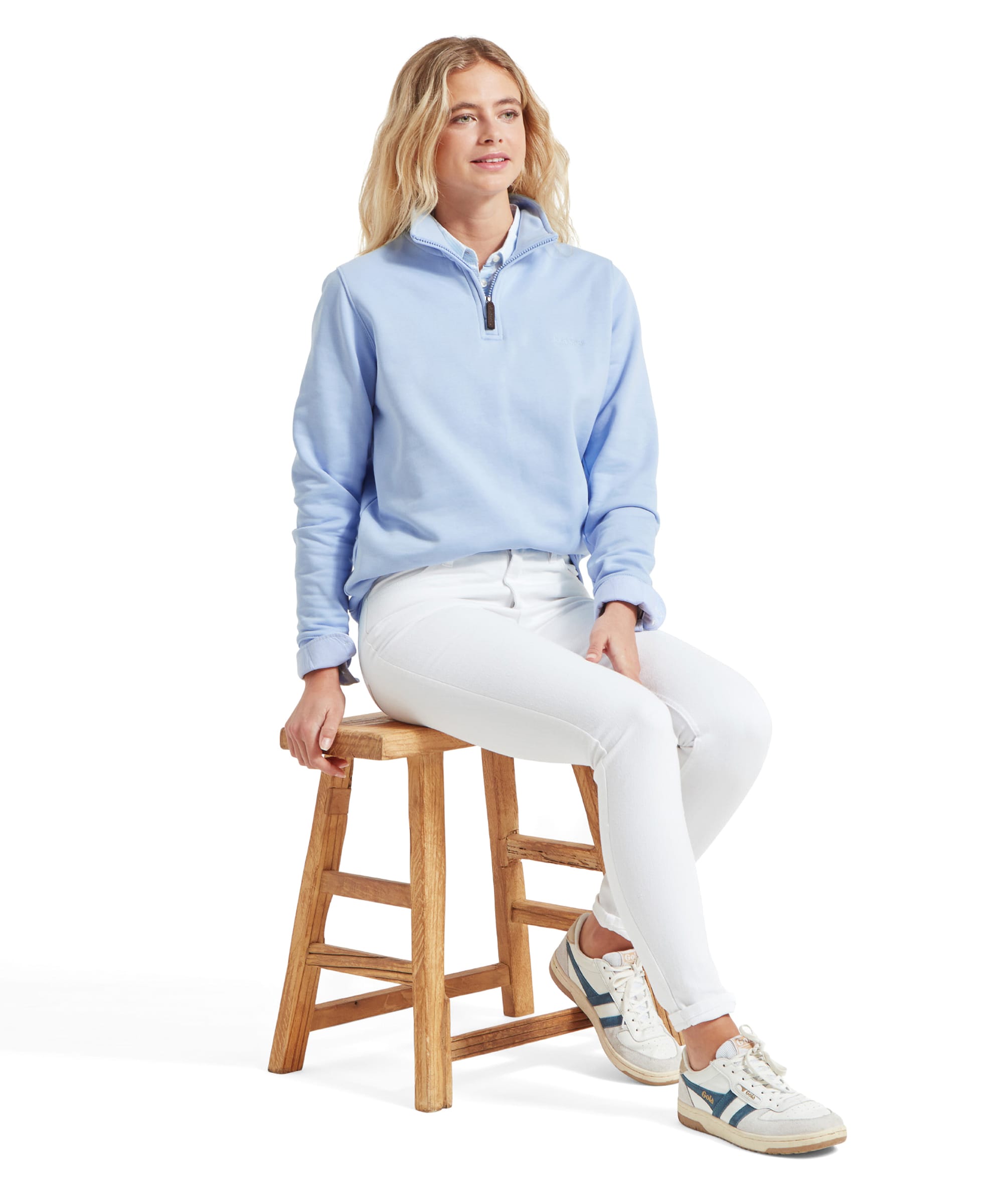 A full-body shot of a woman sitting on a wooden stool, wearing Schöffel Poppy Jean for Womens in White and a light blue sweatshirt with rolled sleeves. She is also wearing white trainers with blue accents.