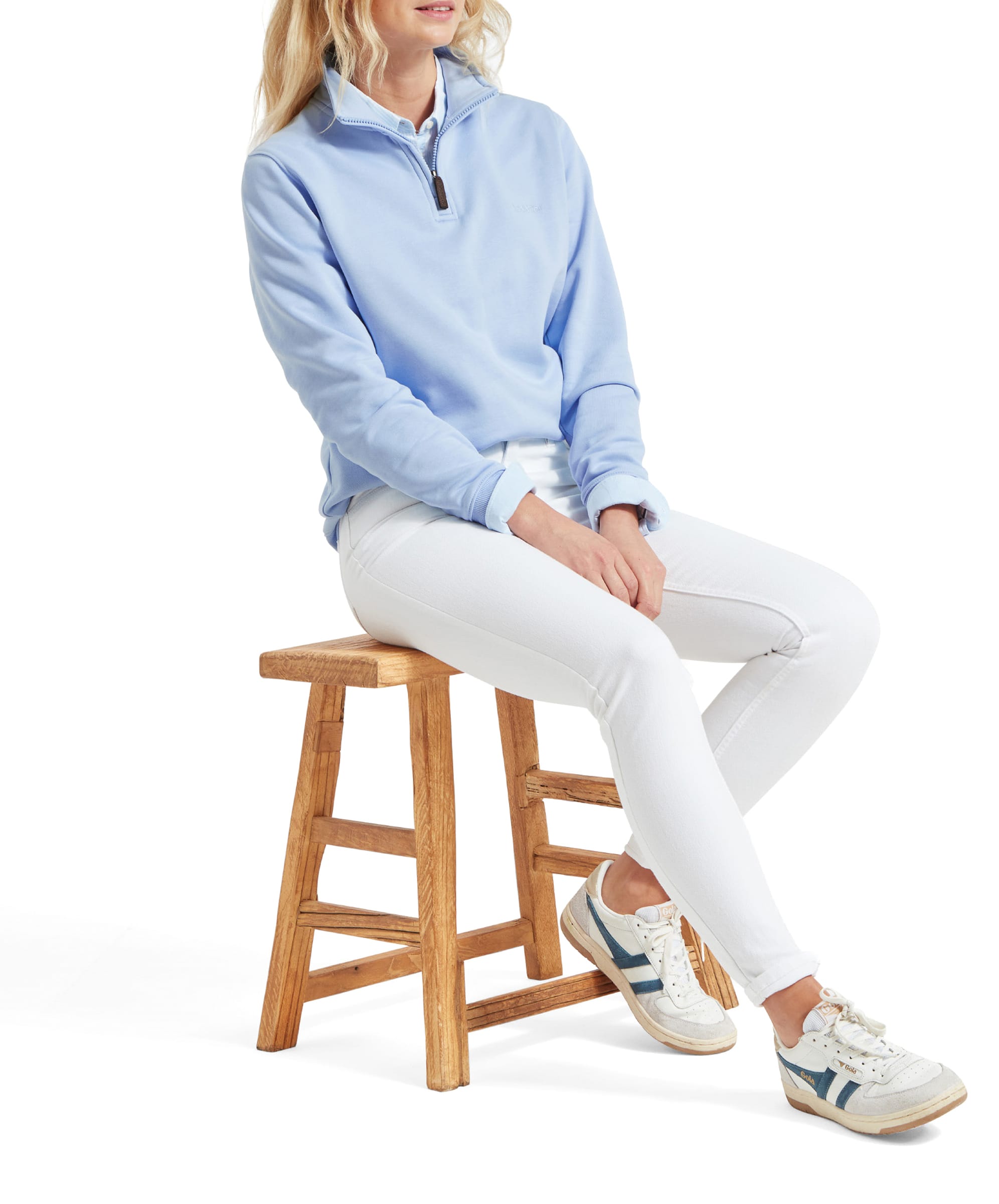 Side view of a woman sitting on a wooden stool, showcasing her Schöffel Poppy Jean for Womens in White and light blue sweatshirt. The jeans have a sleek fit with a slight taper at the ankle, paired with white trainers.