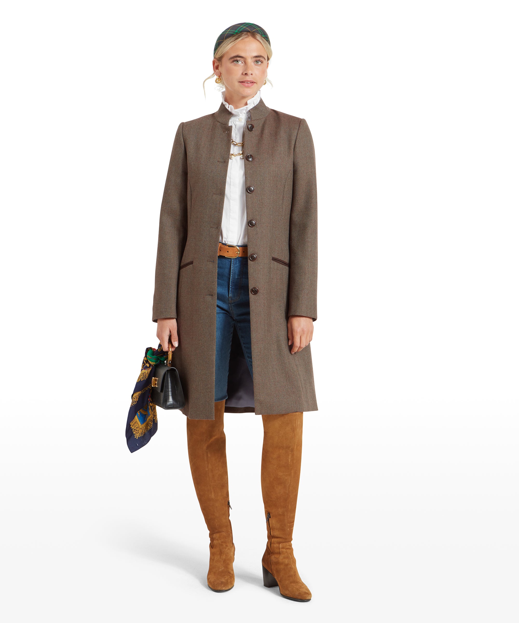 Woman in a Schöffel Portree Tweed Jacket for Women in Green and tan knee-high boots, holding a scarf and a small black handbag. She stands with a neutral expression.