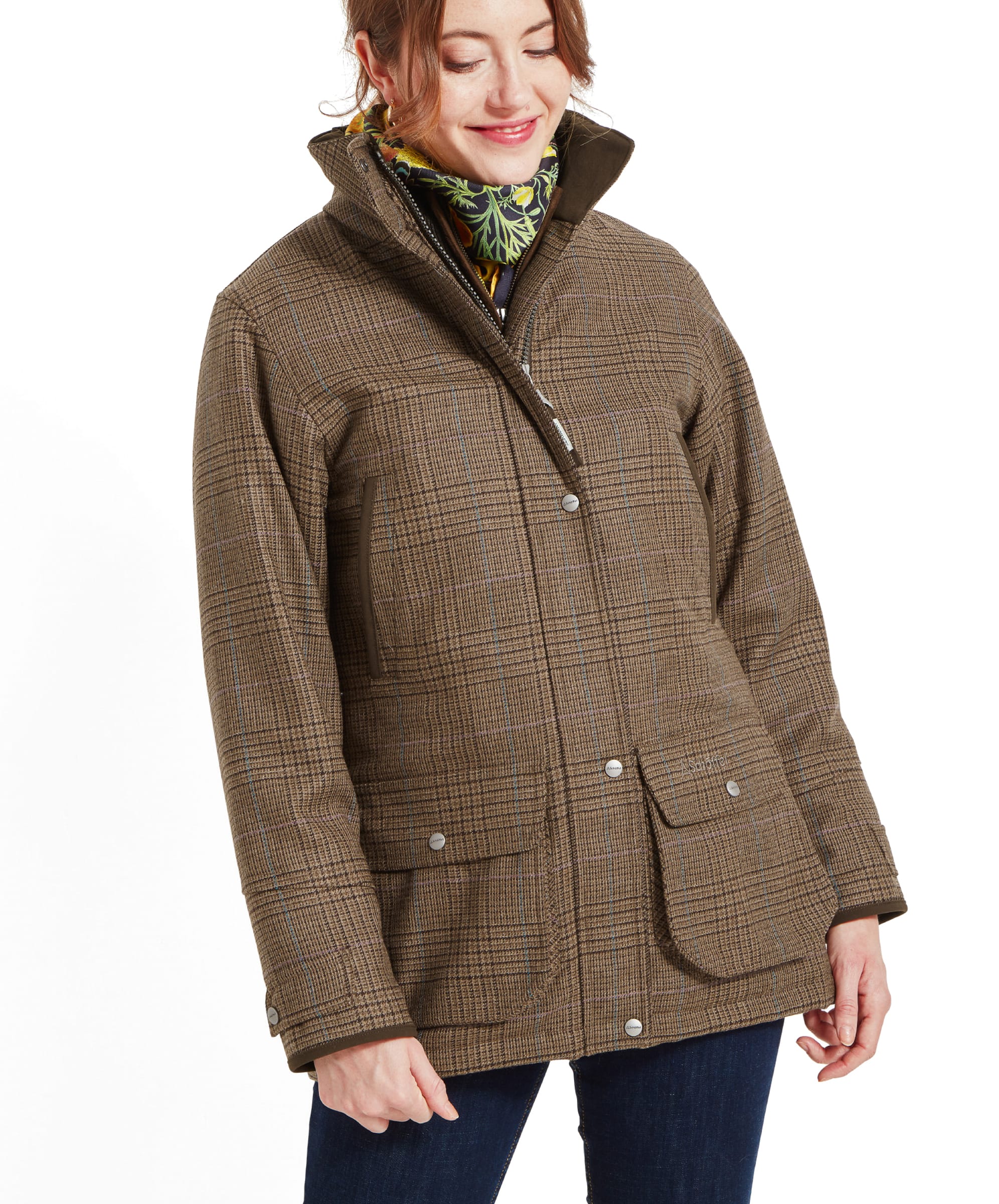 A woman wearing a Schöffel Ptarmigan Tweed Coat for Women in Brown with a high collar and front pockets. She has a slight smile and is accessorised with a floral scarf. The coat has a classic country style with a subtle check pattern.