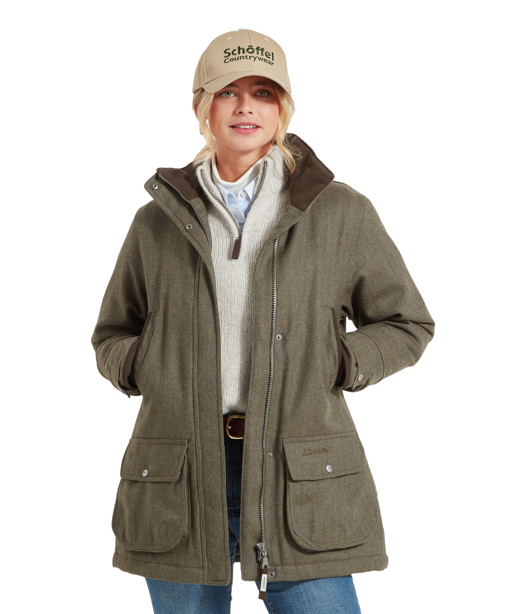 A woman is wearing a Schöffel Ptarmigan Tweed Coat for Women in Green with a brown collar, layered over a light beige sweater and a light blue button-up shirt. She has her hands in the coat pockets and is smiling slightly. The outfit is completed with a beige Schöffel Countrywear baseball cap and blue jeans. The coat features large front pockets with button closures and a visible zipper.