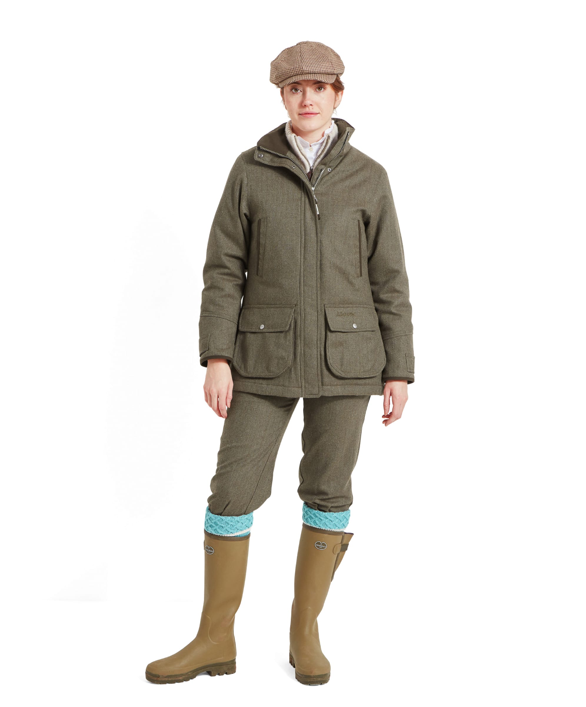 A woman is dressed in a Schöffel Ptarmigan Tweed Coat for Women in Green paired with matching trousers and a beige flat cap. She is wearing tall green rubber boots with blue knitted socks peeking out from the top. The outfit exudes a classic country style, ideal for outdoor activities.