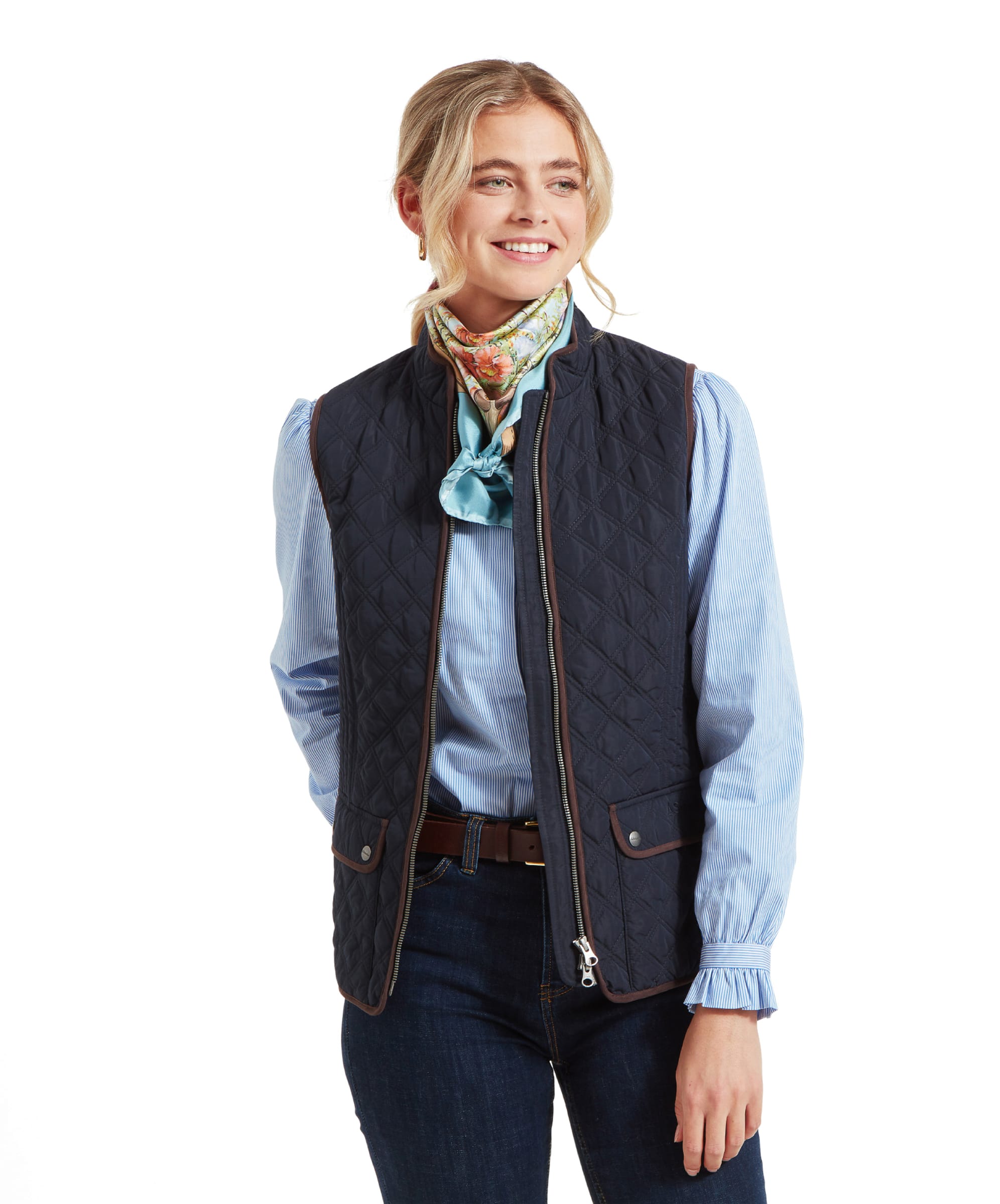 A woman wearing a Schöffel Quilt Gilet for Women in Blue over a light blue shirt, smiling looking away with a hand in her back pocket. She pairs the outfit with dark jeans and a brown belt.