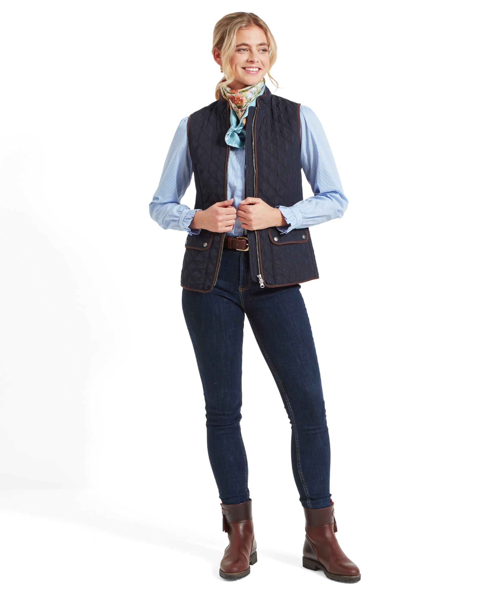 A woman wearing a Schöffel Quilt Gilet for Women in Blue over a light blue shirt, smiling while standing with her hands relaxed in front of her. She pairs the outfit with dark jeans and brown ankle boots.