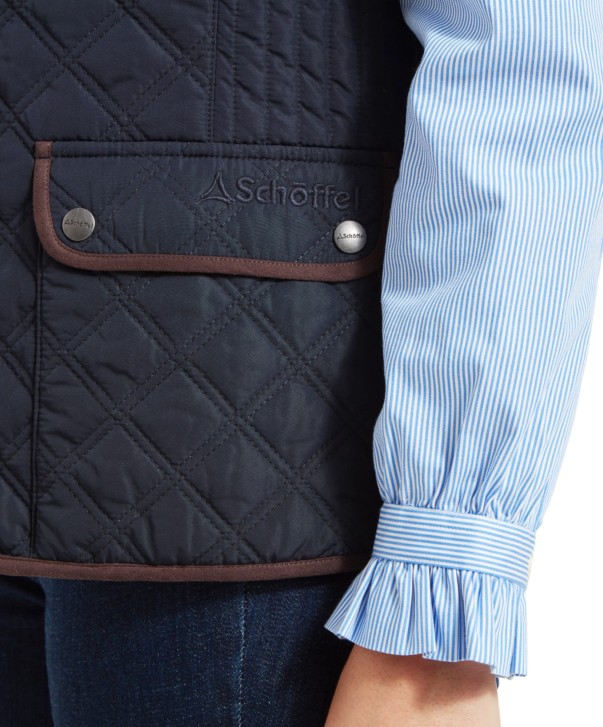 A close-up view of the front pocket of a Schöffel Quilt Gilet for Women in Blue. The pocket has a flap with snap buttons and a brown trim, featuring the brand logo "Schöffel" embroidered above it.