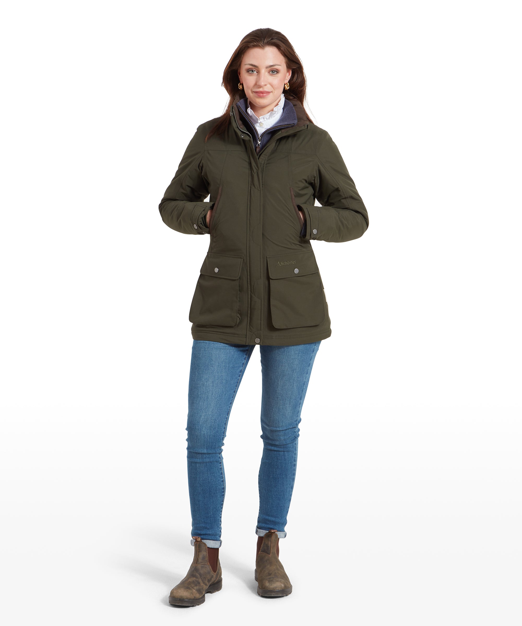 The same woman is standing in the Schöffel Rockingham II Coat for Women in Green, with her hands in the pockets, paired with blue jeans and brown boots, facing the camera.