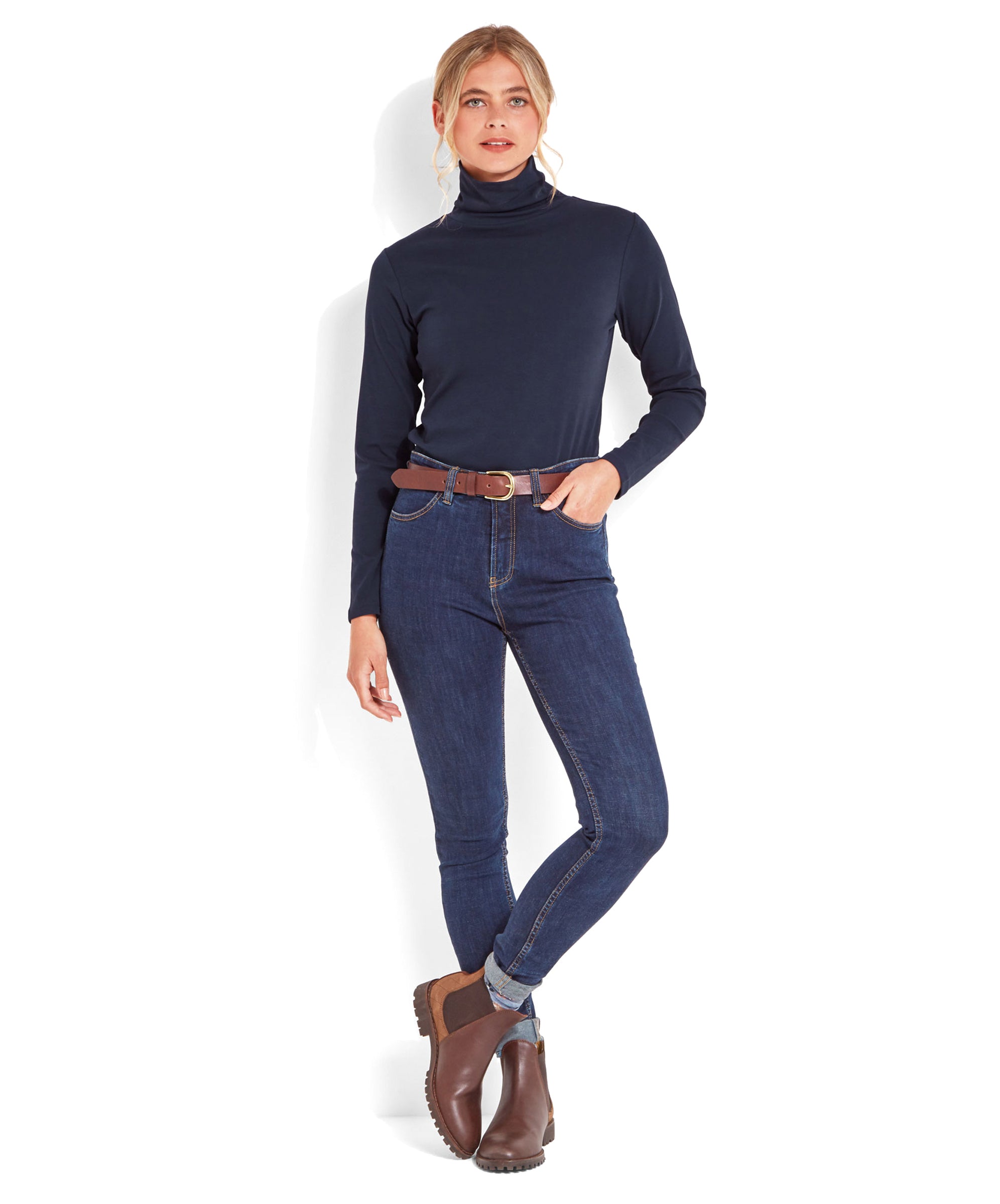 Full-body view of the woman in the Schöffel Rosedale Roll Neck for Women in Navy paired with dark blue jeans and brown boots, standing with one hand in her pocket.