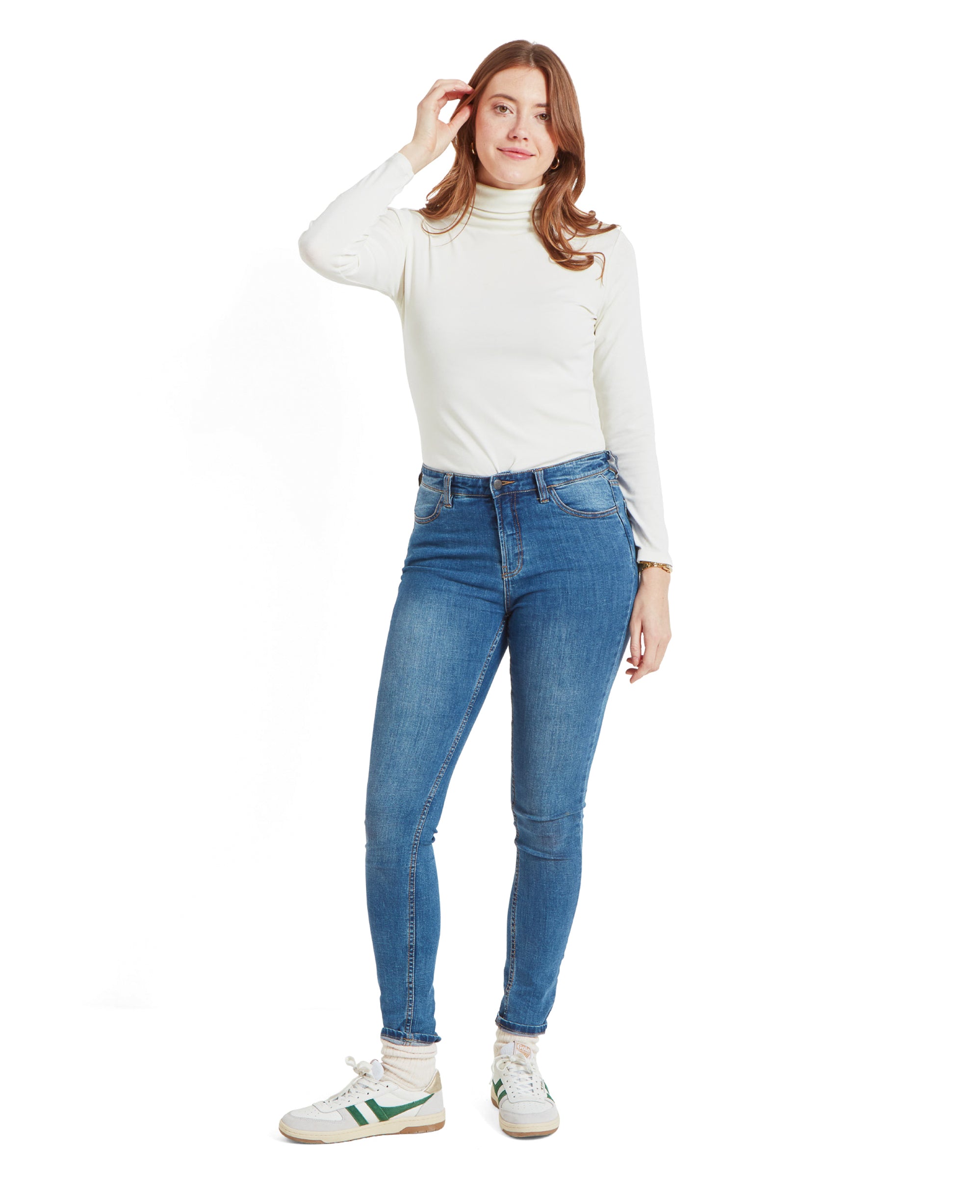 A full-body view of the woman in the Schöffel Rosedale Roll Neck for Women in White, paired with blue jeans and white trainers, standing with one hand touching her hair.