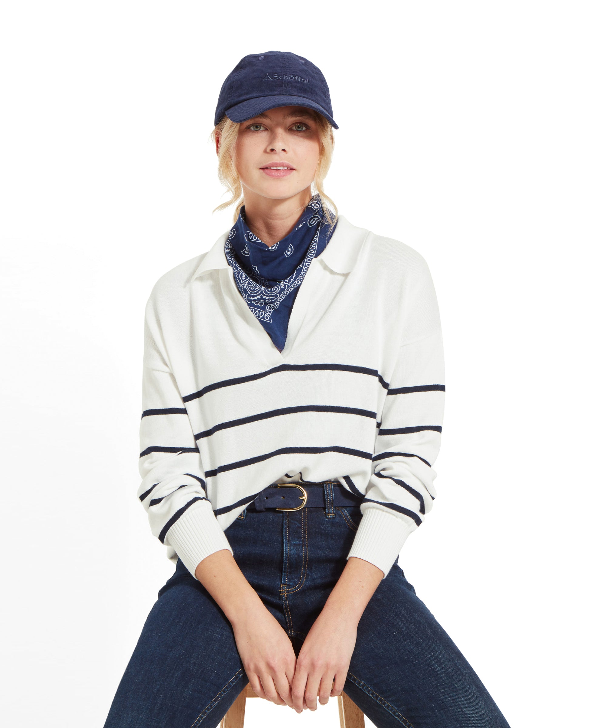 A woman sitting on a stool, wearing a Schöffel Roseland Jumper for Women in Navy, dark blue jeans, a navy blue cap, and a navy blue bandana around her neck.