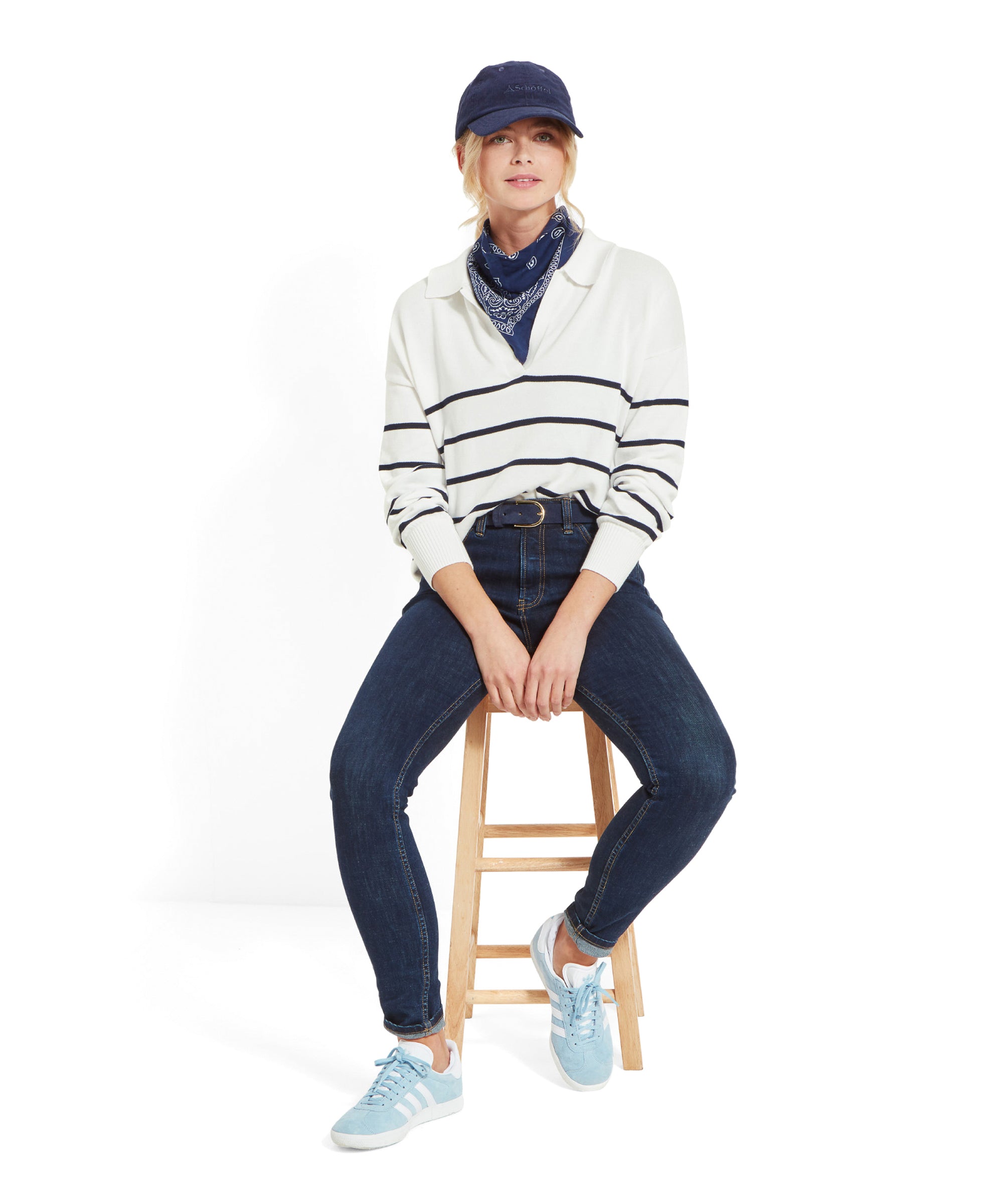 A woman sitting on a stool, a full-body view wearing a Schöffel Roseland Jumper for Women in Navy, dark blue jeans, light blue trainers, a navy blue cap, and a navy blue bandana around her neck.