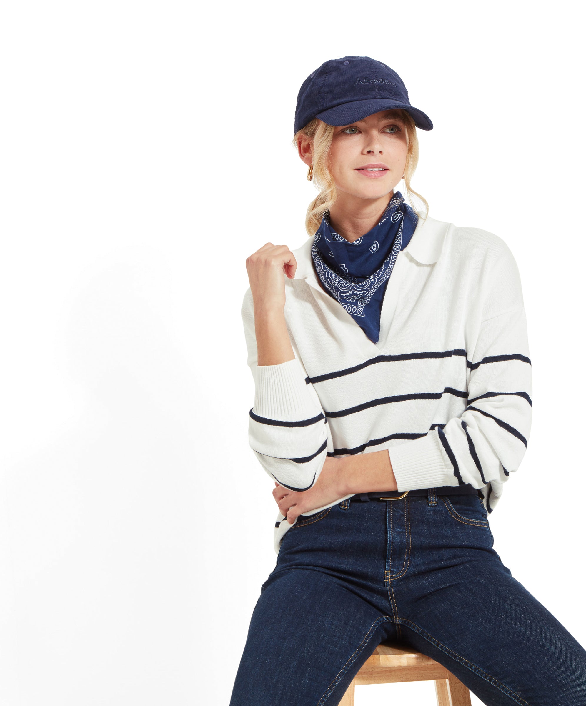 A woman sitting on a stool, wearing a Schöffel Roseland Jumper for Women in Navy, dark blue jeans, light blue trainers, a navy blue cap, and a navy blue bandana around her neck, with her hand resting on her chin.