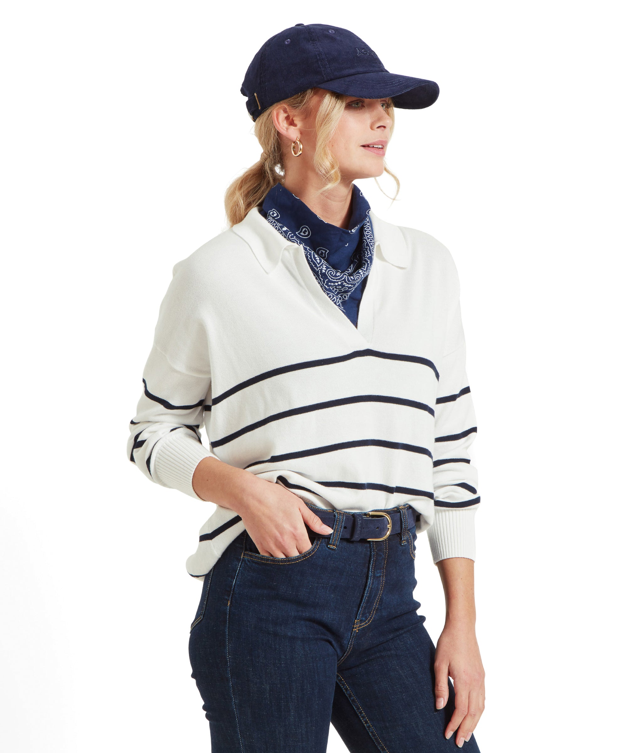 A woman wearing a Schöffel Roseland Jumper for Women in Navy, dark blue jeans, light blue trainers, a navy blue cap, and a navy blue bandana around her neck, standing sideways and looking to the left.