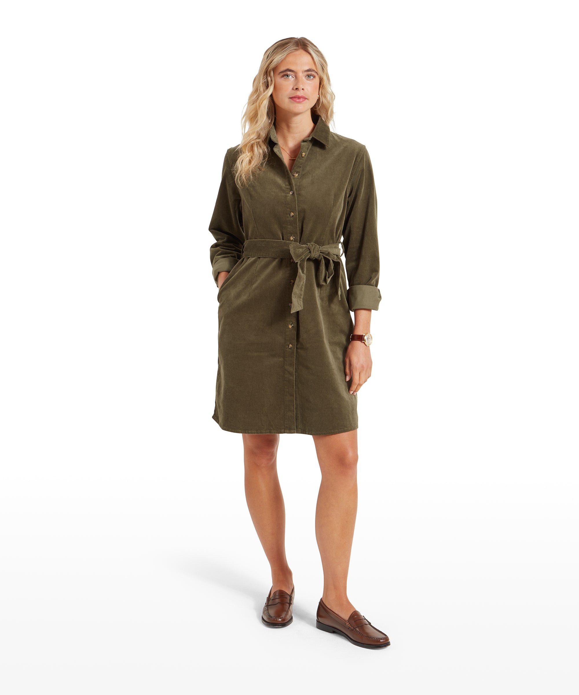 A woman is wearing a Schöffel Sage Cord Dress for Womens in Green with long sleeves. The dress is belted at the waist and features button-down front details. She is standing with her hands in her pockets, looking slightly to the side with a relaxed expression.