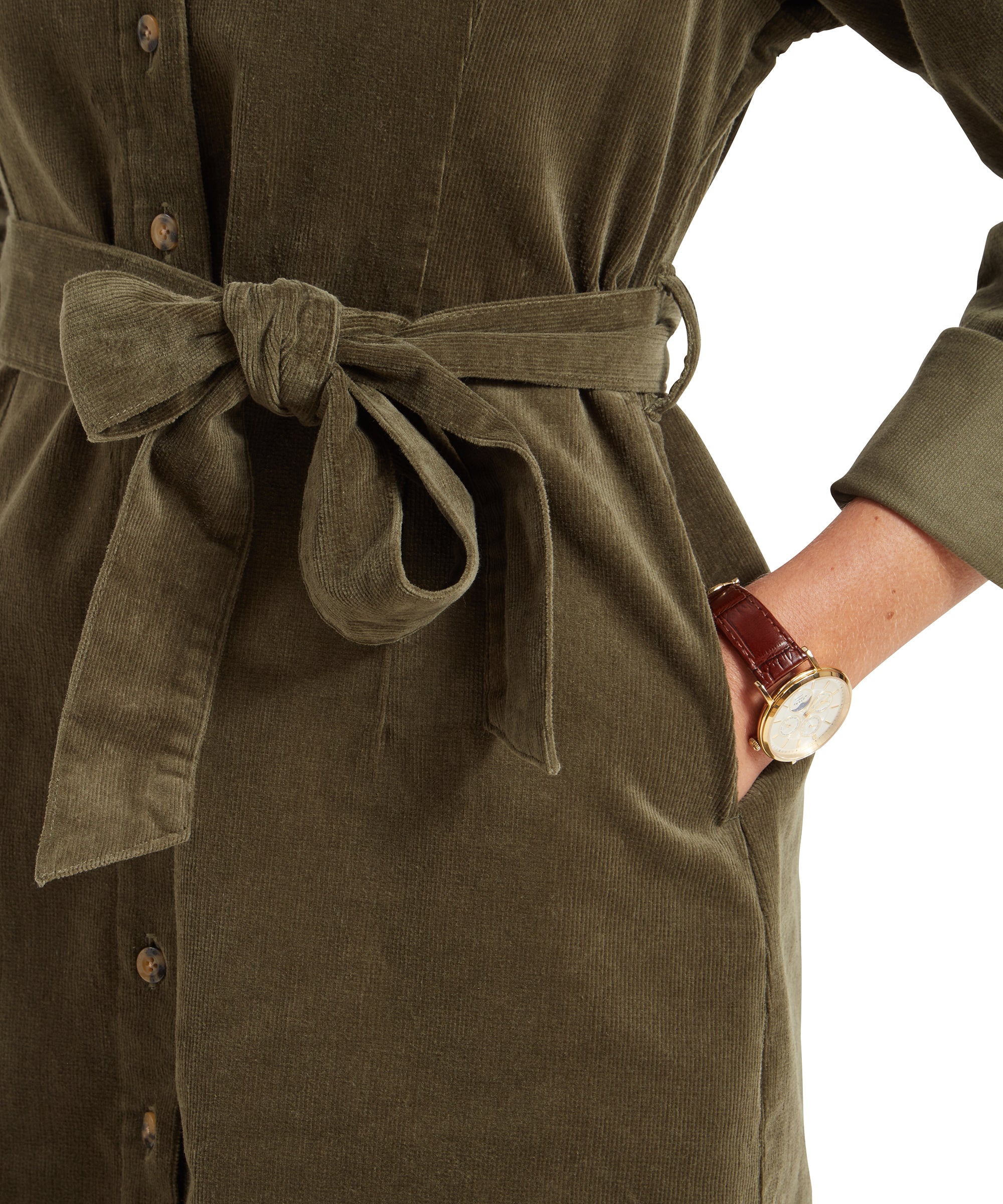 Close-up view of the Schöffel Sage Cord Dress for Womens in Green's waist area, highlighting the tied belt and the texture of the corduroy fabric. The woman's hand is in the dress pocket, and she is wearing a wristwatch with a brown leather strap.