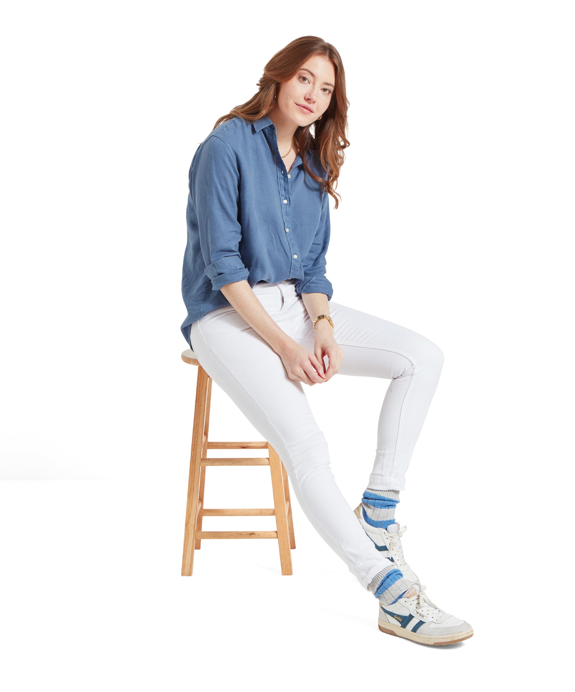 Full-body shot of the woman sitting on a stool, wearing a Schöffel Salthouse Linen Shirt for Women in Blue, white jeans, and white trainers.