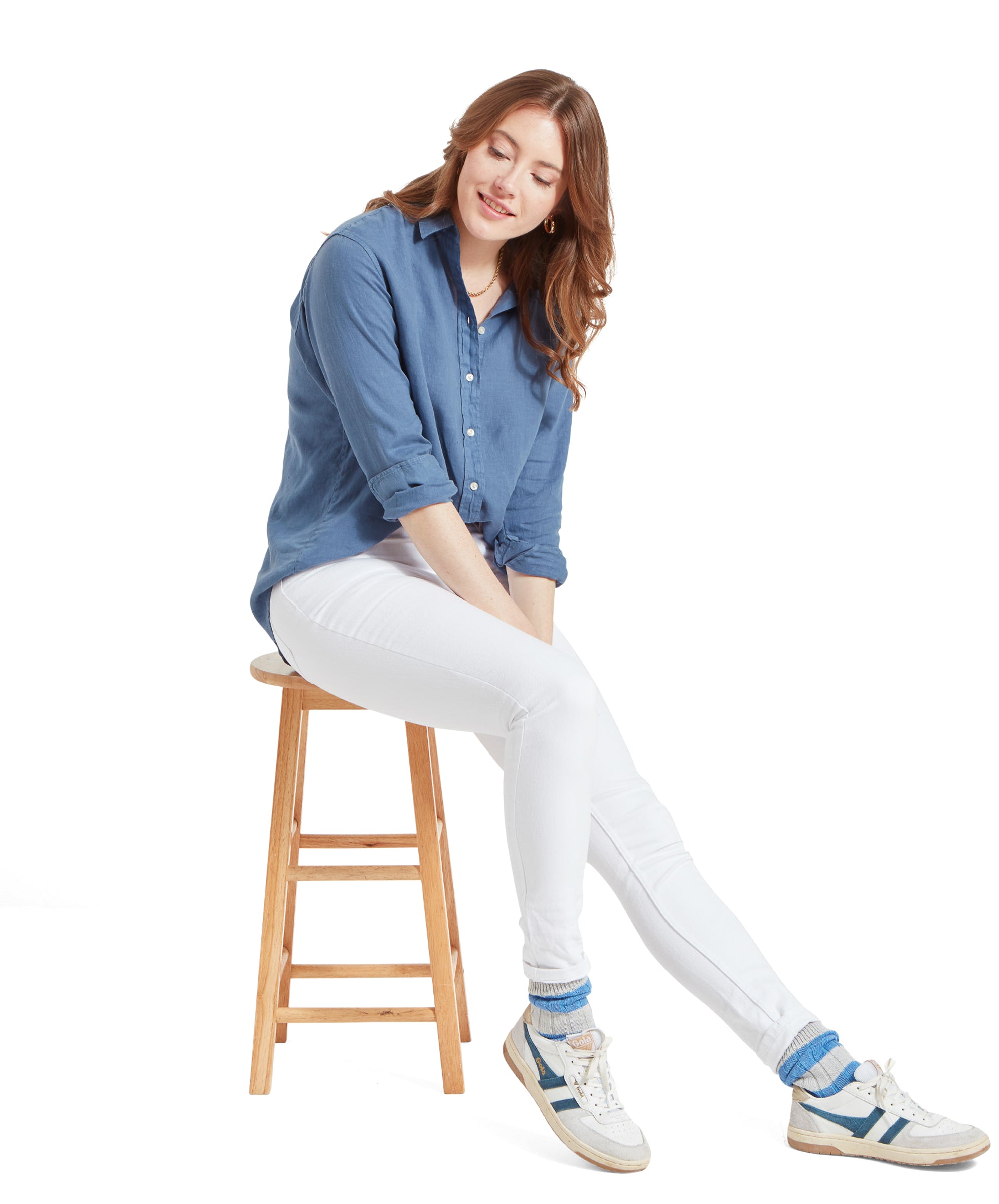 Woman seated on a stool, smiling and looking down, wearing a Schöffel Salthouse Linen Shirt for Women in Blue, white jeans, and white trainsers.