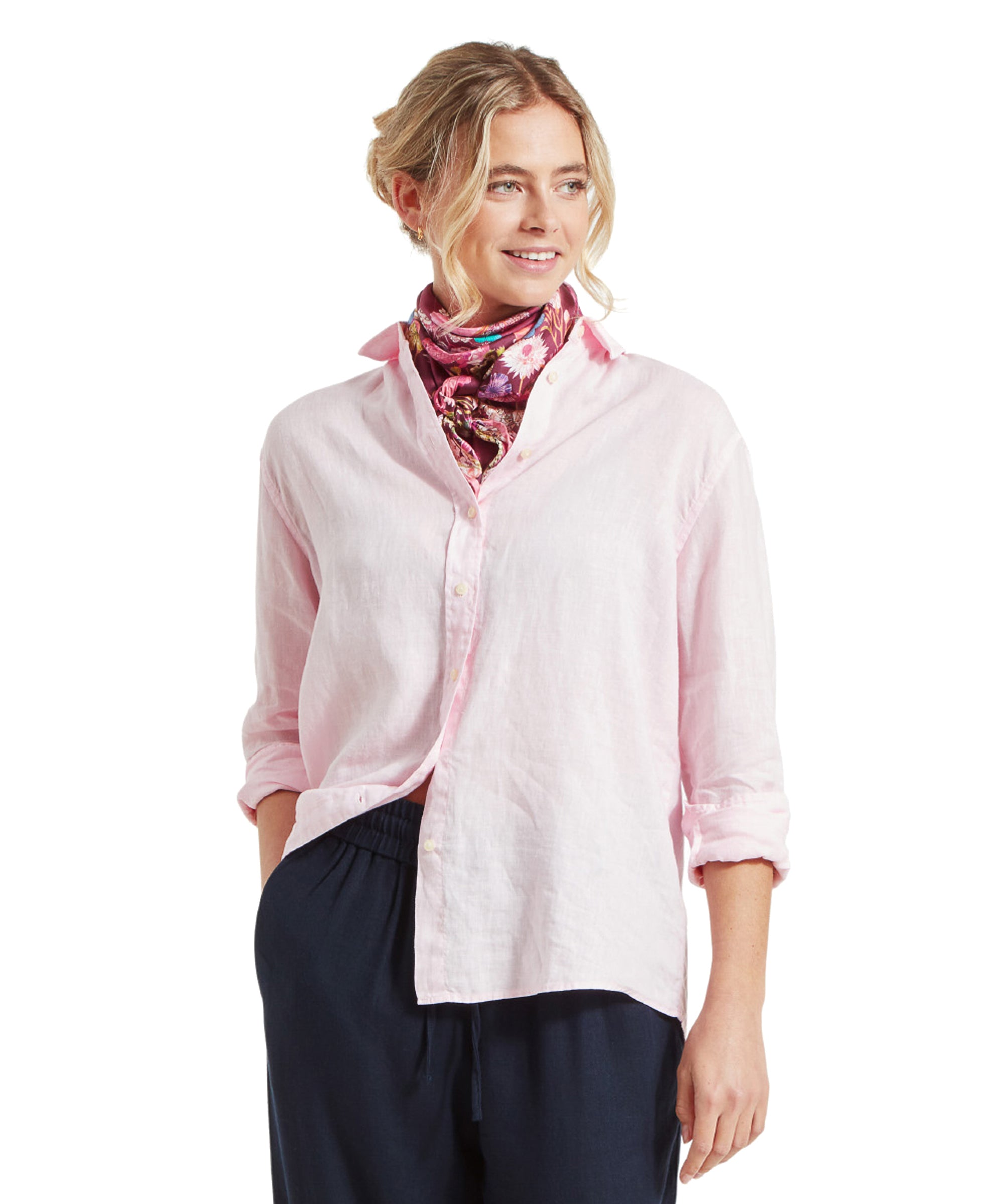 A woman is wearing a Schöffel Salthouse Linen Shirt for Women in Pink paired with a colorful floral scarf around her neck. She has her hands in her pockets and is smiling slightly, looking off to the side. The shirt is buttoned up and has a relaxed fit.