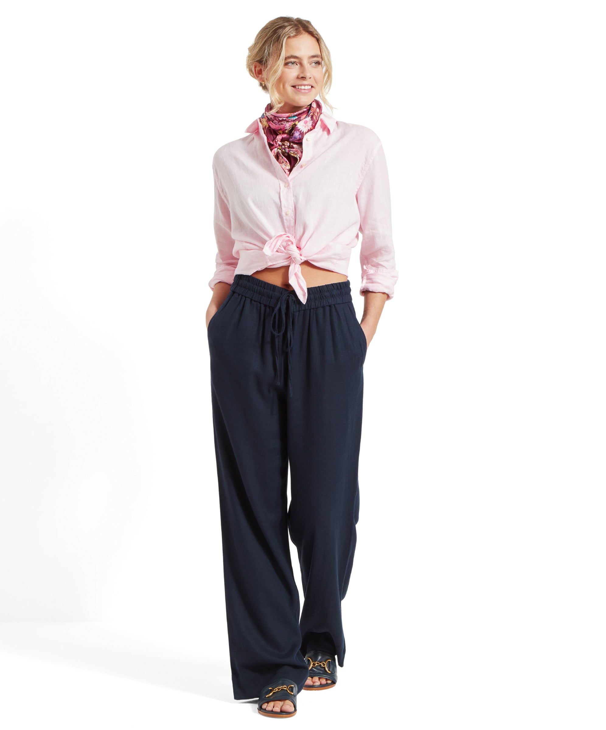 The same woman is shown in a full-body shot, with the Schöffel Salthouse Linen Shirt for Women in Pink tied at the waist, revealing her midriff. She is wearing loose, dark blue jeans with an elastic waist and black sandals. She is smiling and looking to the side.