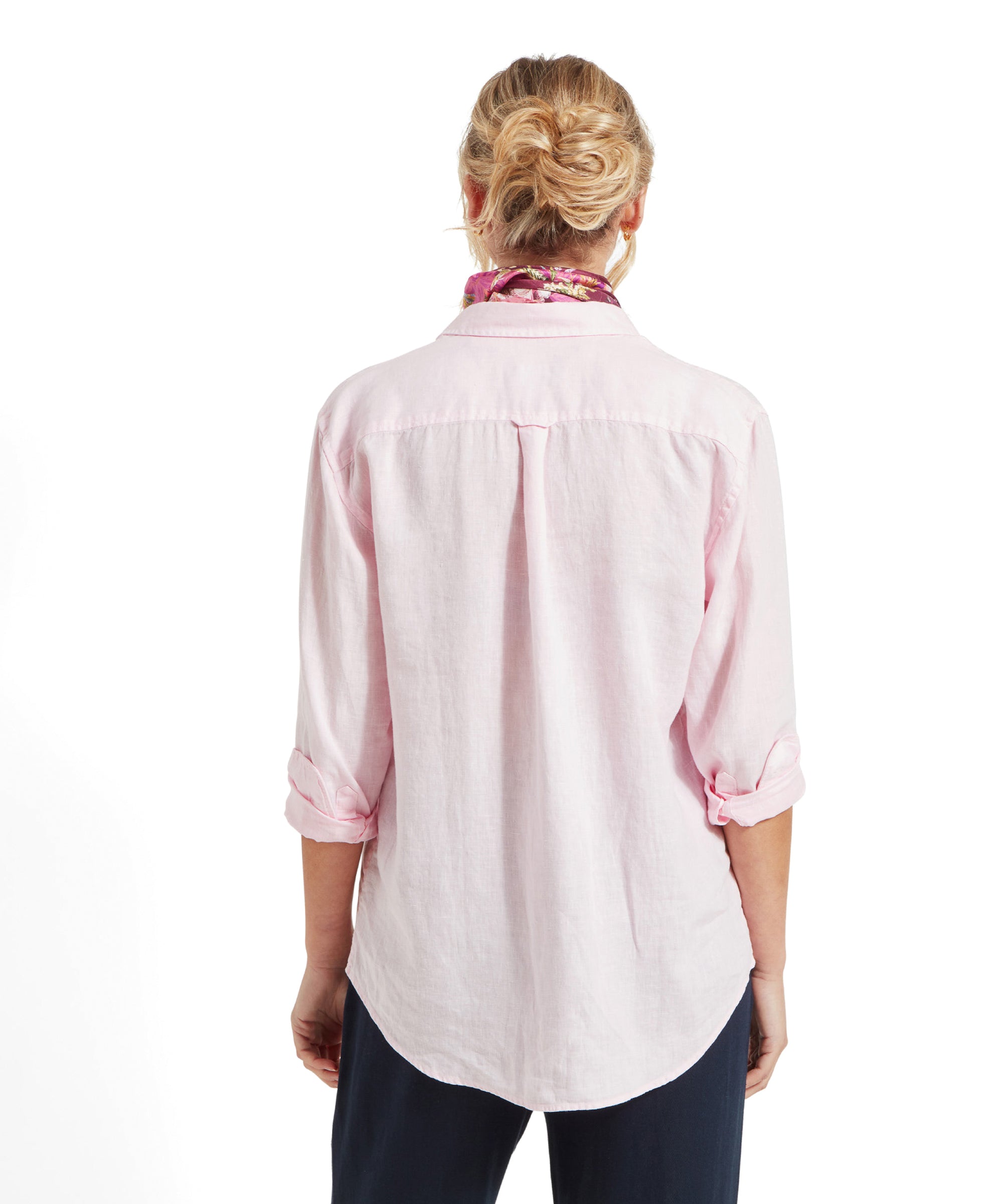 The woman is seen from the back, wearing the Schöffel Salthouse Linen Shirt for Women in Pink. The shirt is long and covers her hips. Her hair is styled in a loose bun, and the colorful floral scarf is visible around her neck.