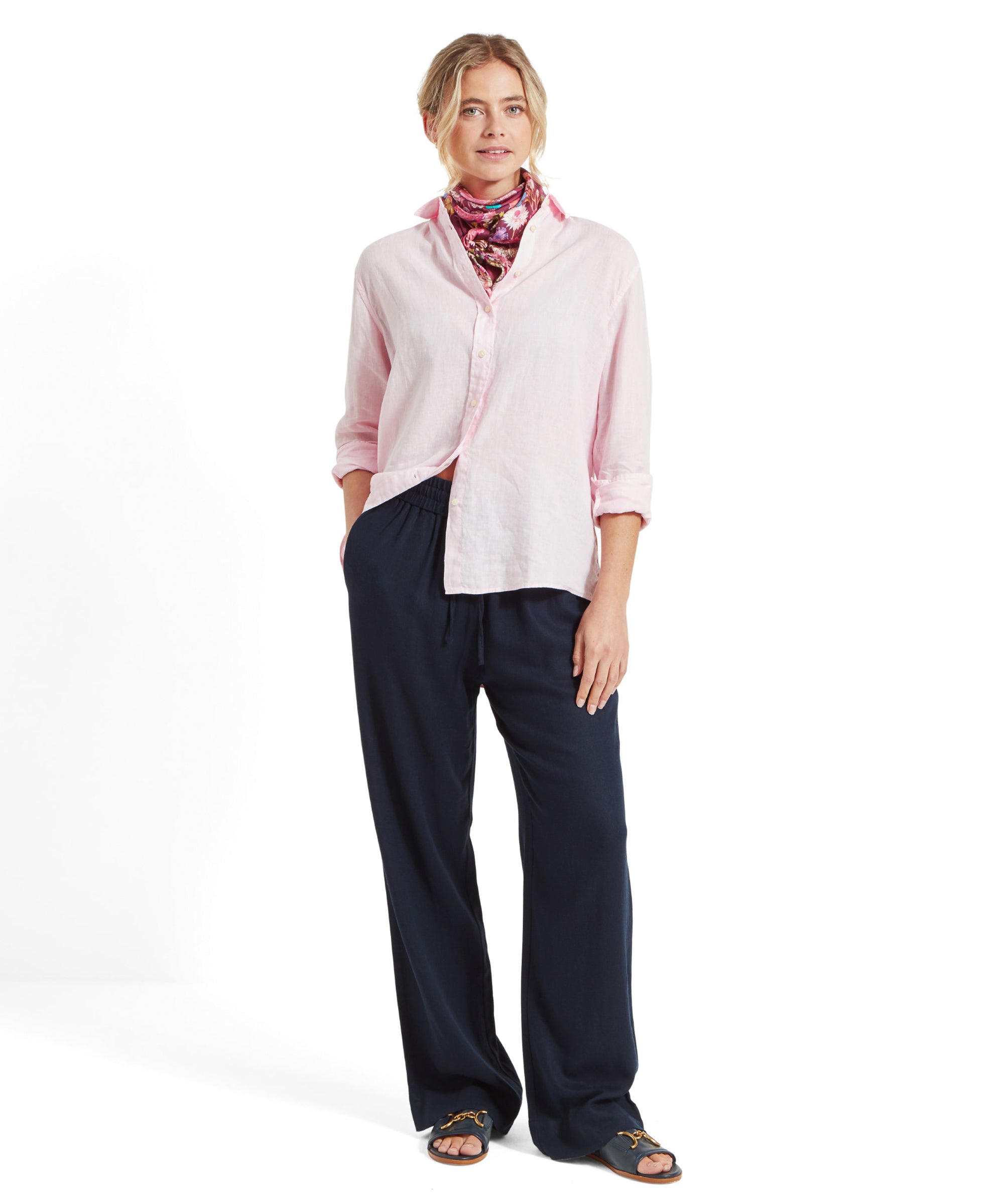 The same woman is shown in a full-body shot, with the Schöffel Salthouse Linen Shirt for Women in Pink slightly pulled tpo the side with the models hand in her pocket. She is wearing loose, dark blue jeans with an elastic waist and black sandals. She is smiling and looking forward.