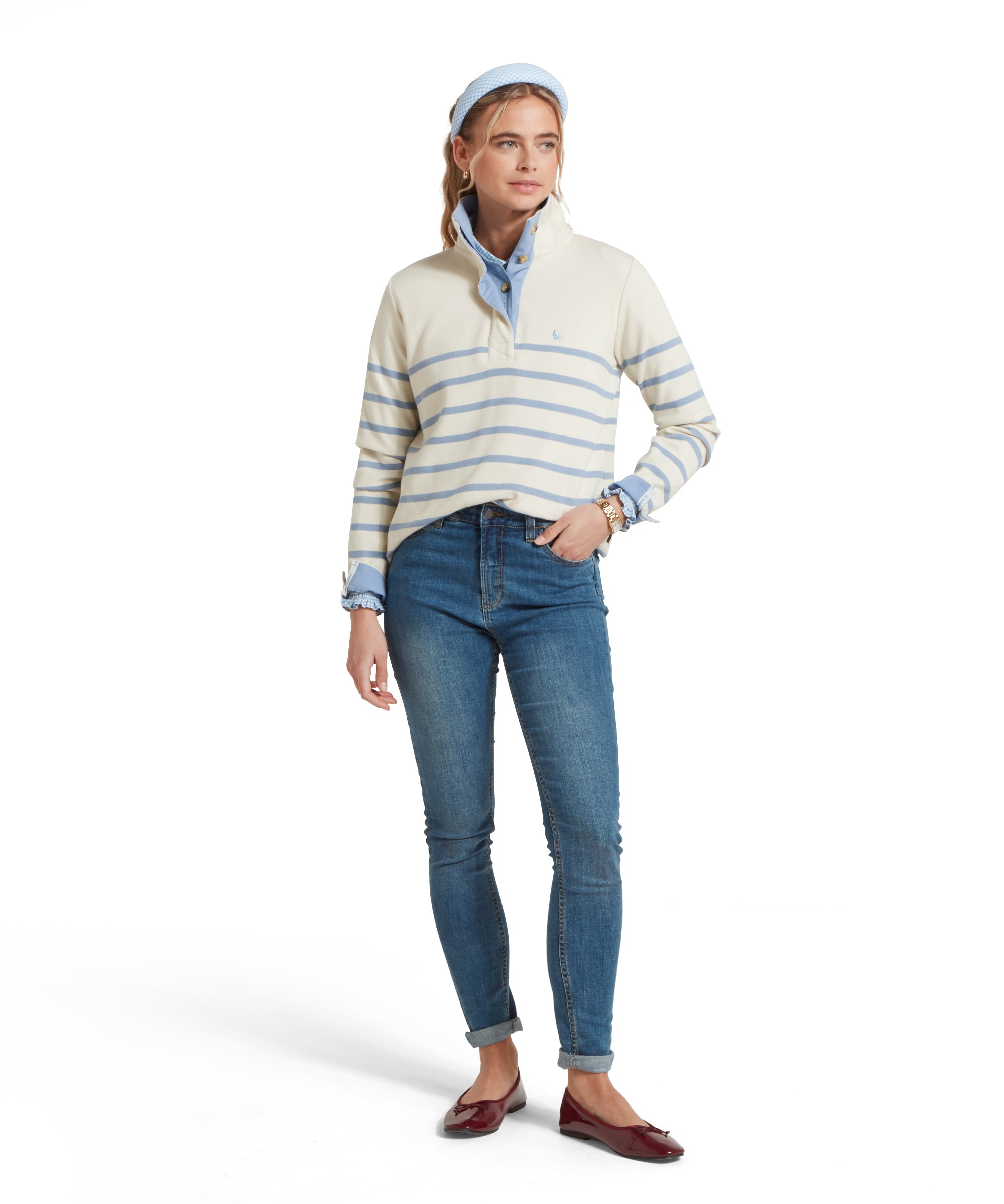 Female model standing in a relaxed pose, wearing a beige and blue striped Schöffel sweatshirt with a small embroidered bird logo on the chest, blue shirt, denim blue jeans and  dark red pumps.