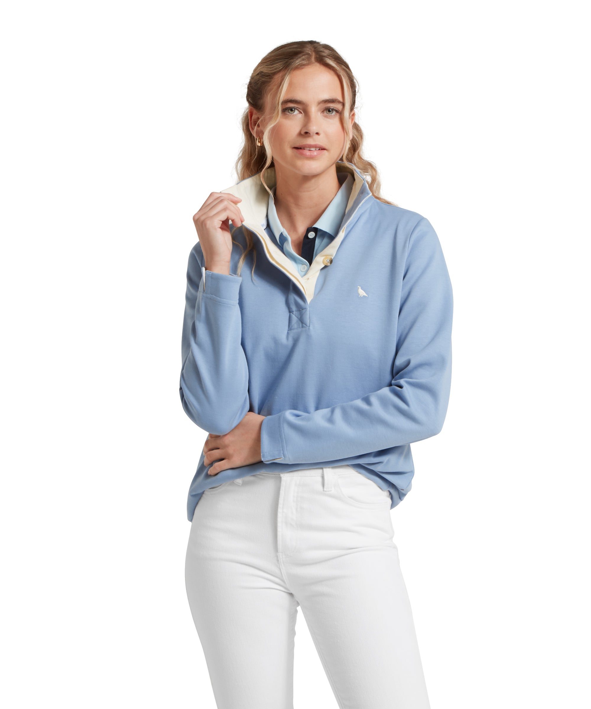 Female model standing in a relaxed pose, wearing a blue Schöffel sweatshirt with a small embroidered bird logo on the chest, blue polo shirt and white jeans.