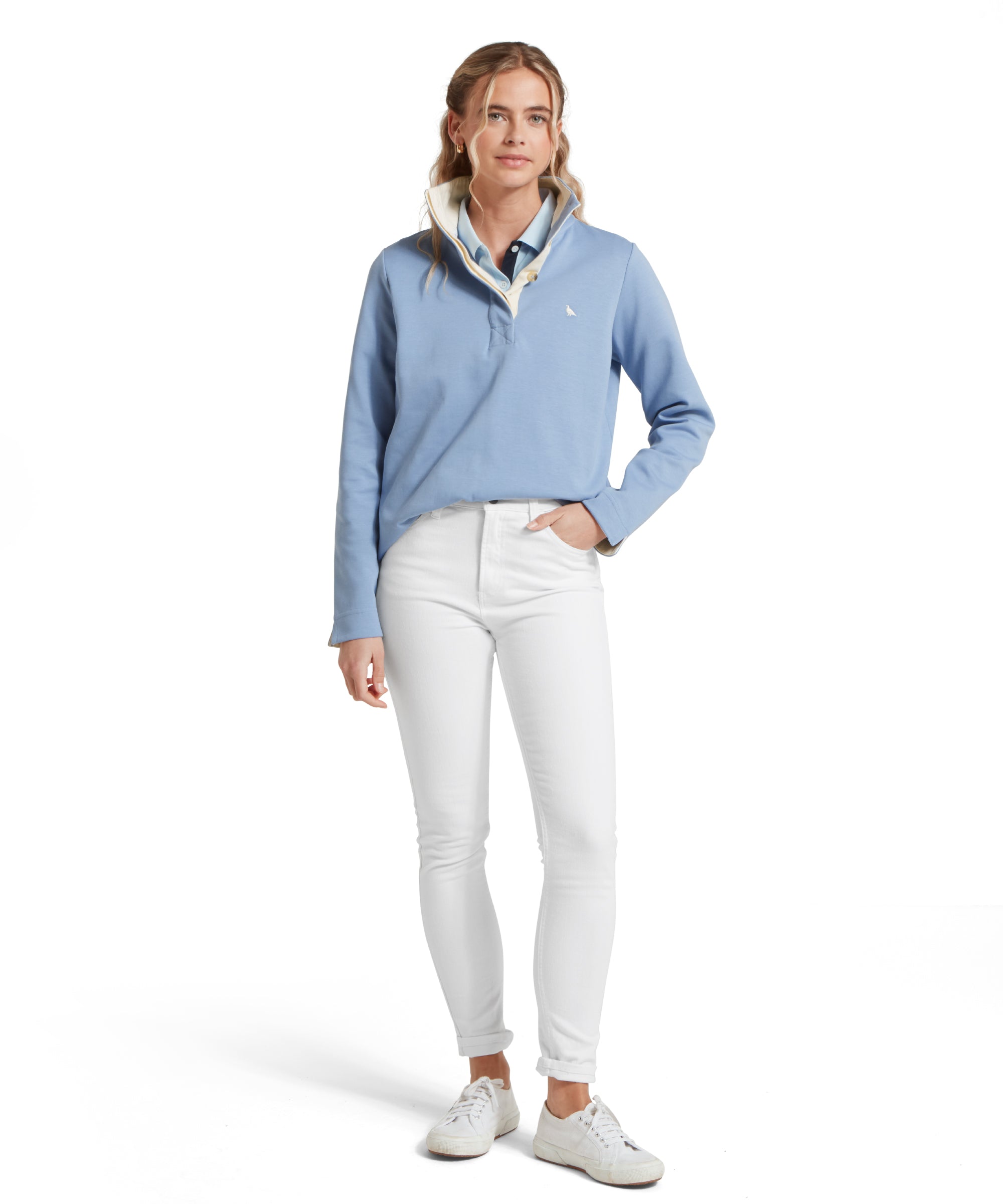 Female model standing in a relaxed pose, wearing a blue Schöffel sweatshirt with a small embroidered bird logo on the chest, blue polo shirt, white jeans and white trainers.