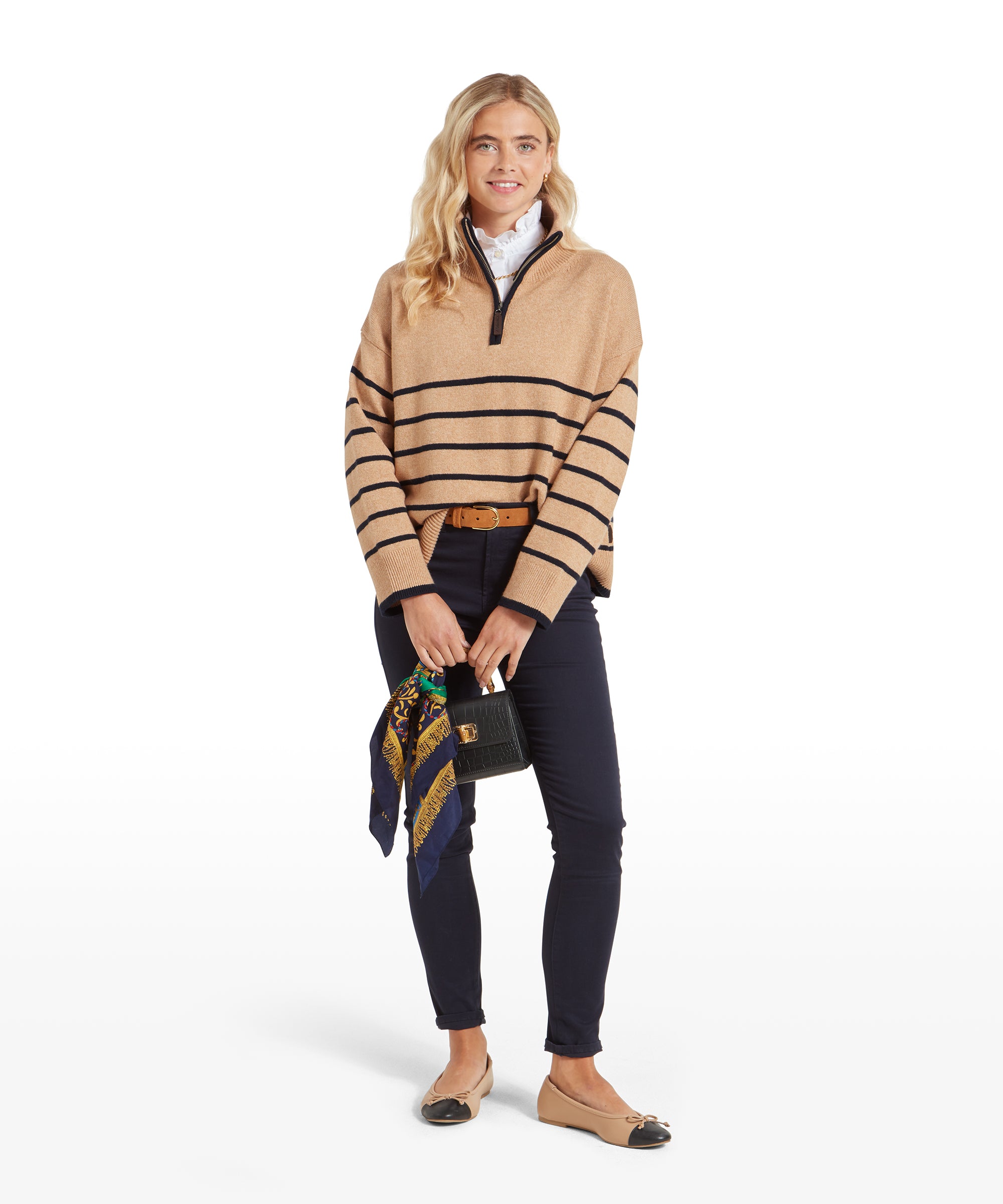 The woman stands holding a small black handbag with a colourful scarf attached. She wears the Schöffel Sandsend 1/4 Jumper for Women in Beige, navy trousers, and tan belt, completing the look with a polished and casual style.