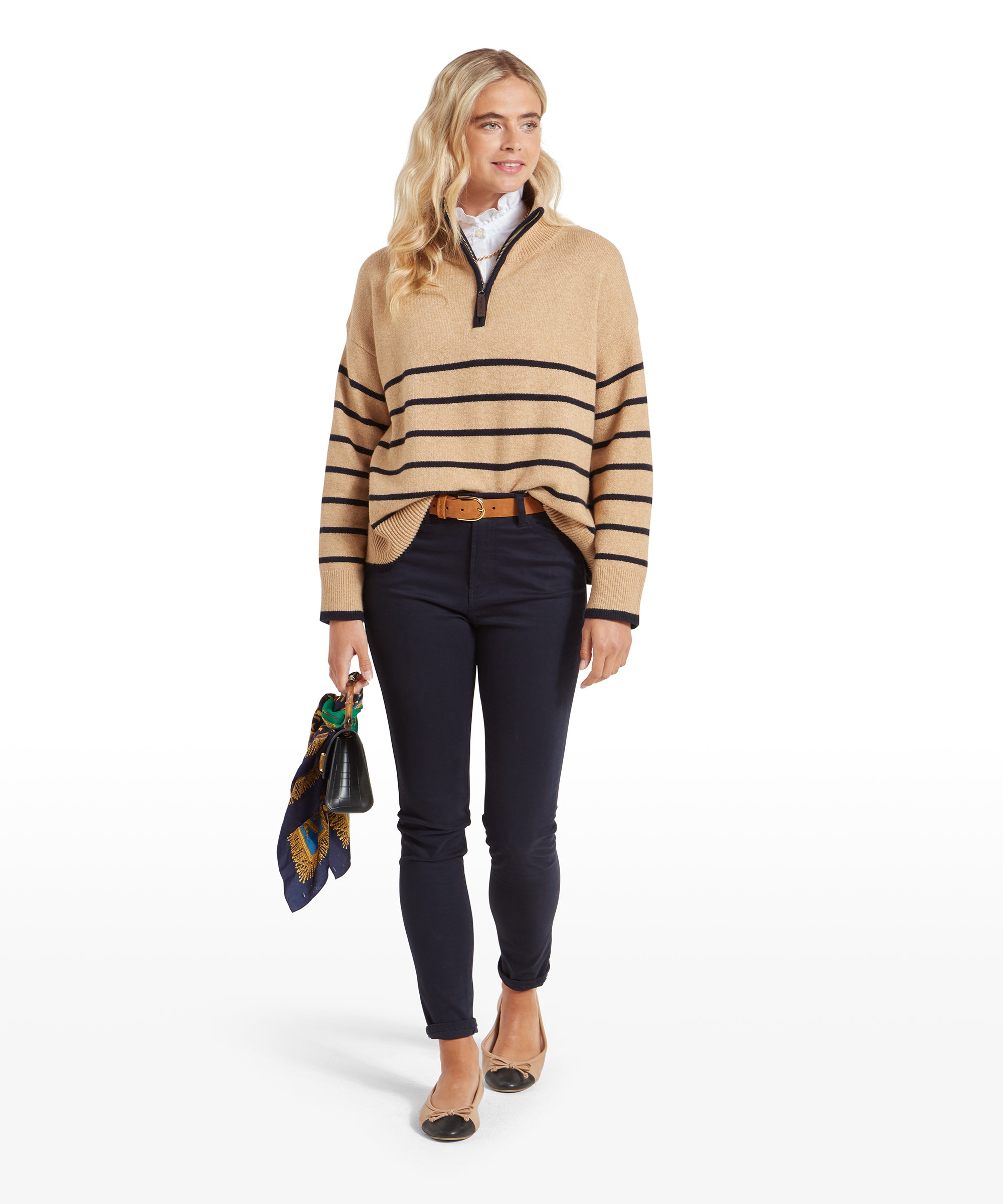 The woman is walking forward, wearing the Schöffel Sandsend 1/4 Jumper for Women in Beige, navy trousers, and tan belt, holding a small black handbag with a scarf, projecting an effortlessly chic appearance.