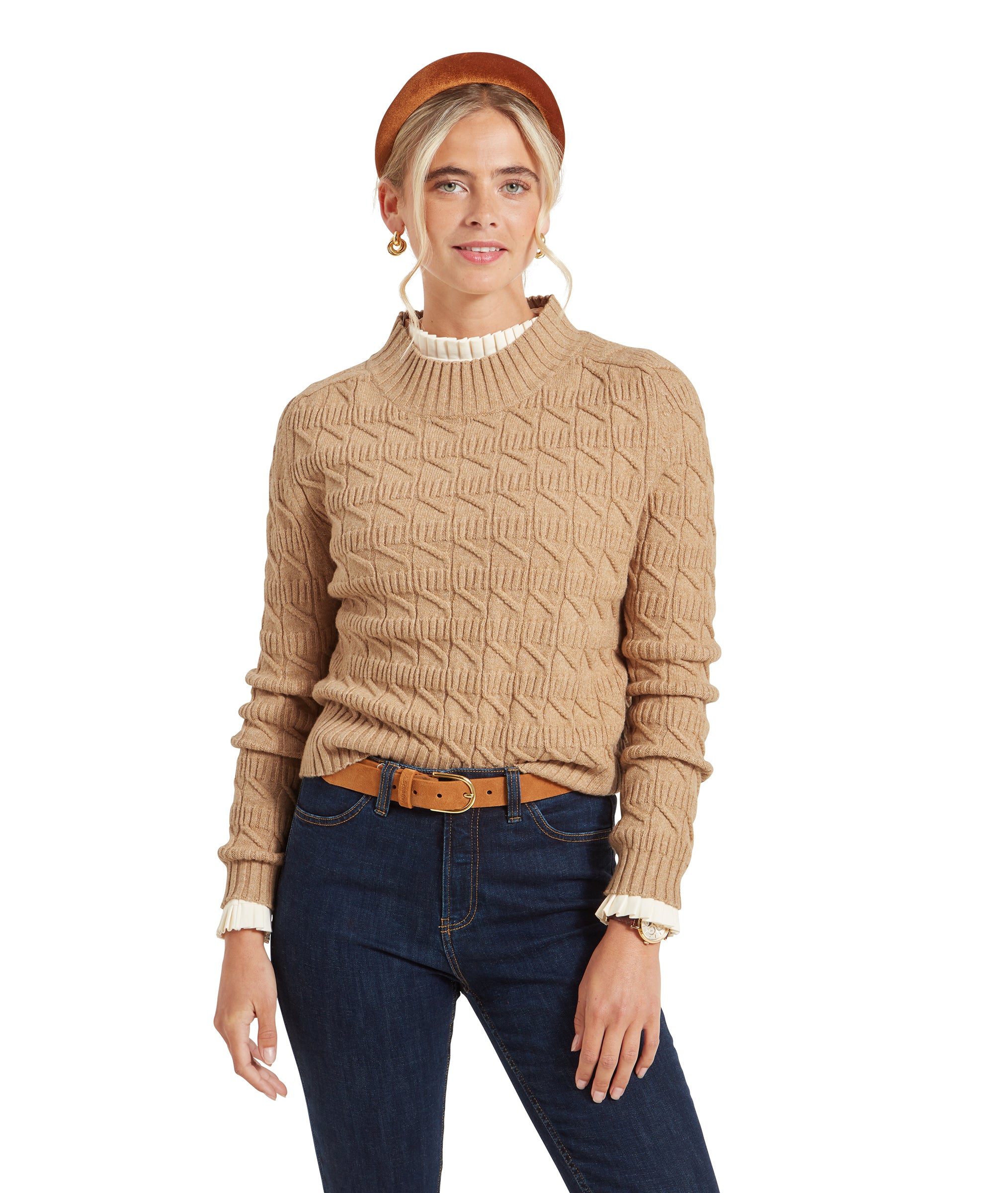 A woman wearing a Schöffel Sandsend Cable Jumper for Women in Beige with a geometric pattern, paired with dark blue jeans. She is standing and smiling, with her hands relaxed at her sides. She is also wearing a brown headband and gold earrings.