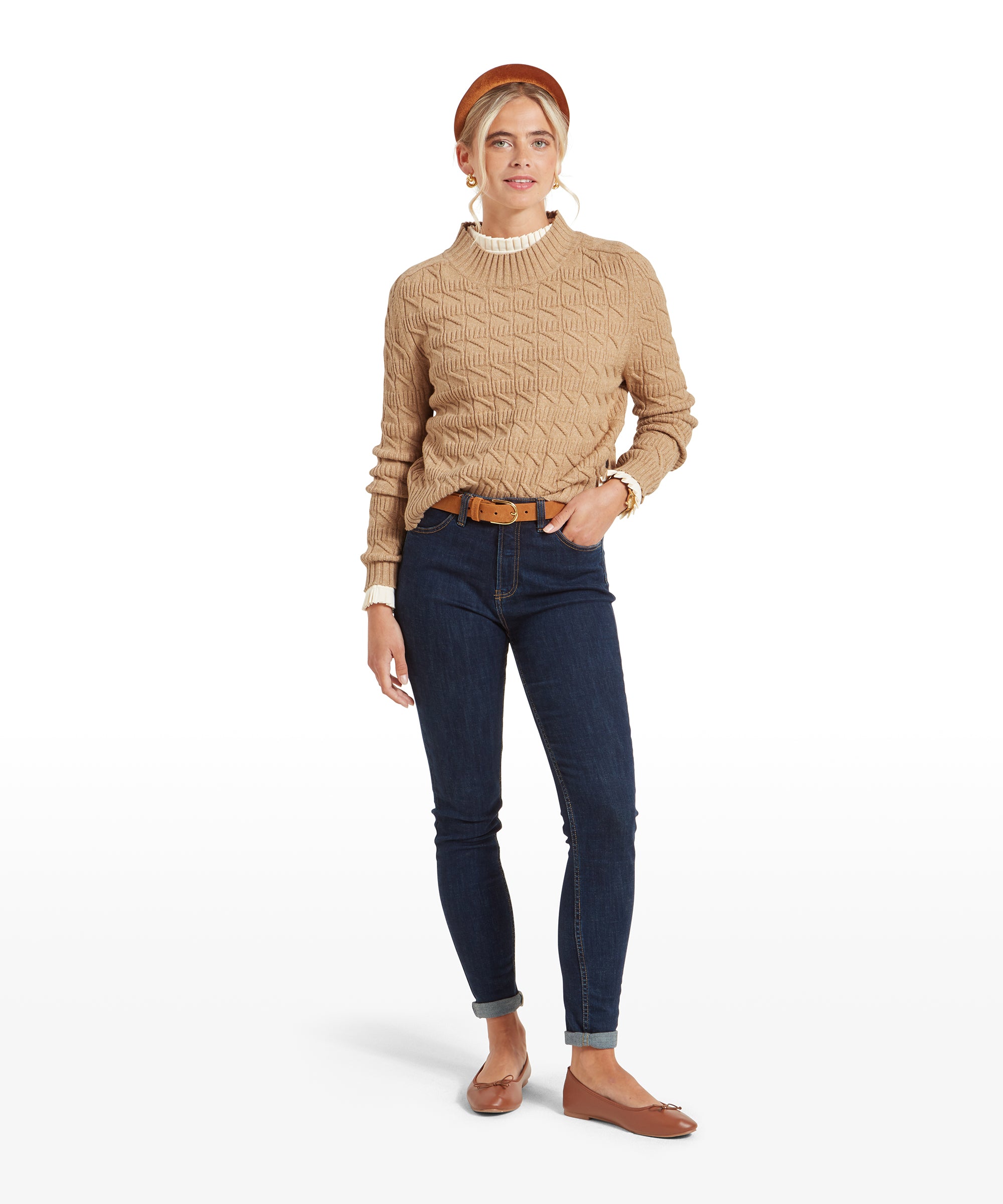 Full-body view of a woman wearing a Schöffel Sandsend Cable Jumper for Women in Beige with a geometric pattern, dark blue jeans, and brown flats. She is standing with a relaxed posture, one hand in her pocket.